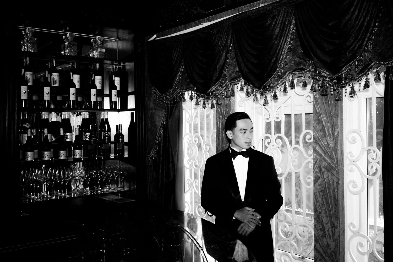 Man in tuxedo standing elegantly by ornate white gate near a bar in black and white photography.