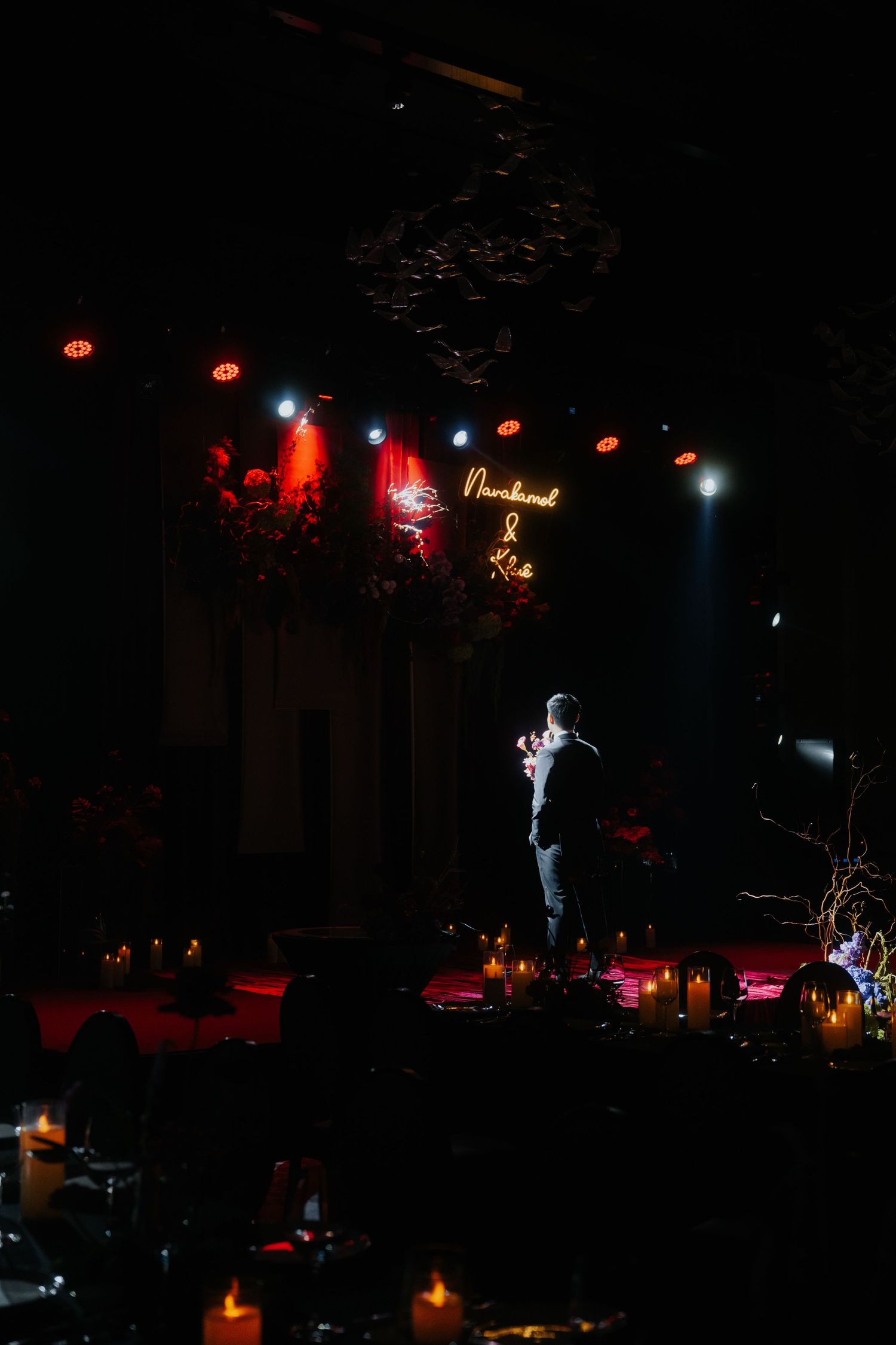 A person stands on a dimly lit stage with candles and a neon sign at an elegant wedding reception.