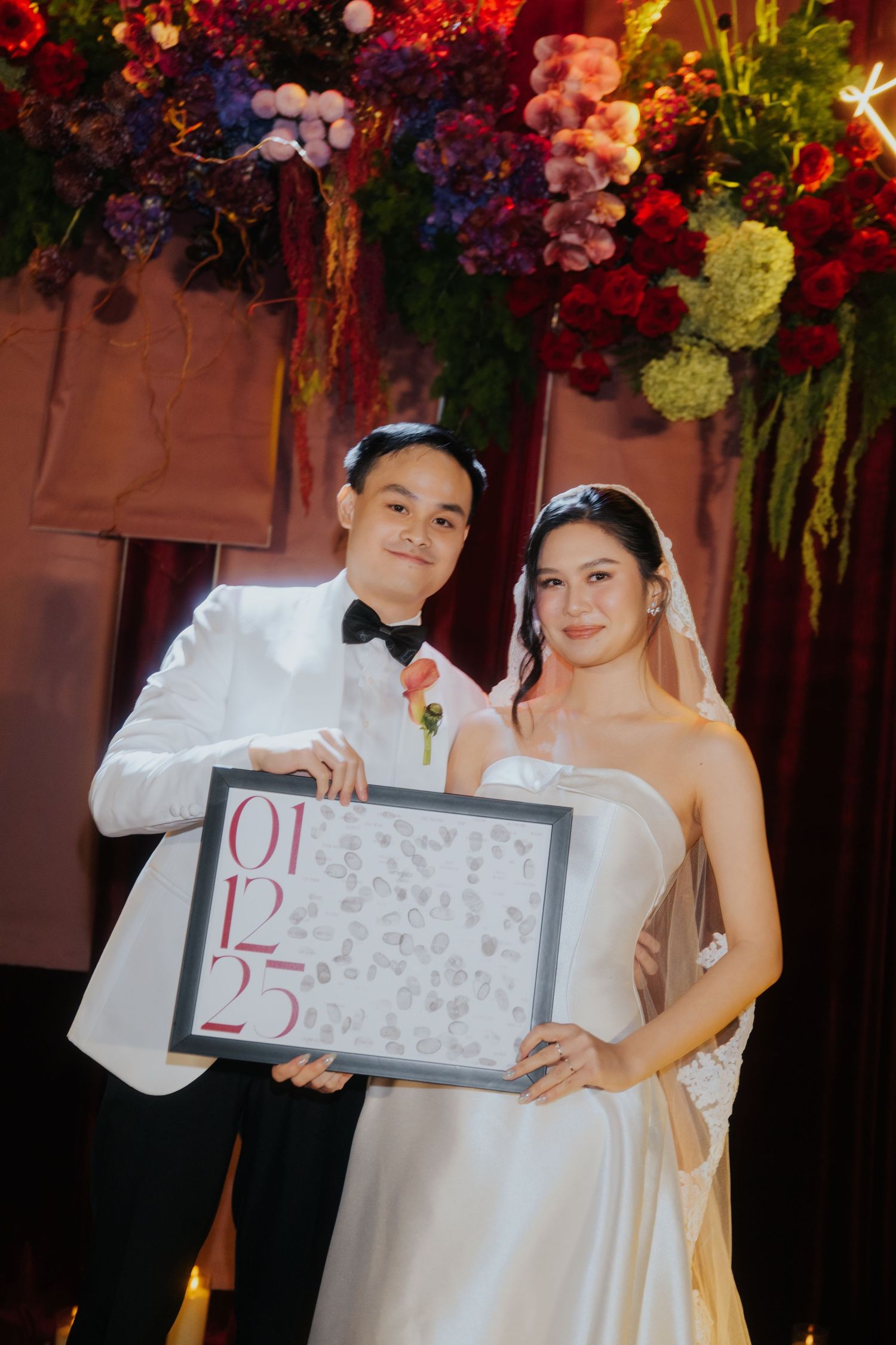 Bride and groom in white attire holding a framed wedding date sign 01.12.25 at elegant floral reception.