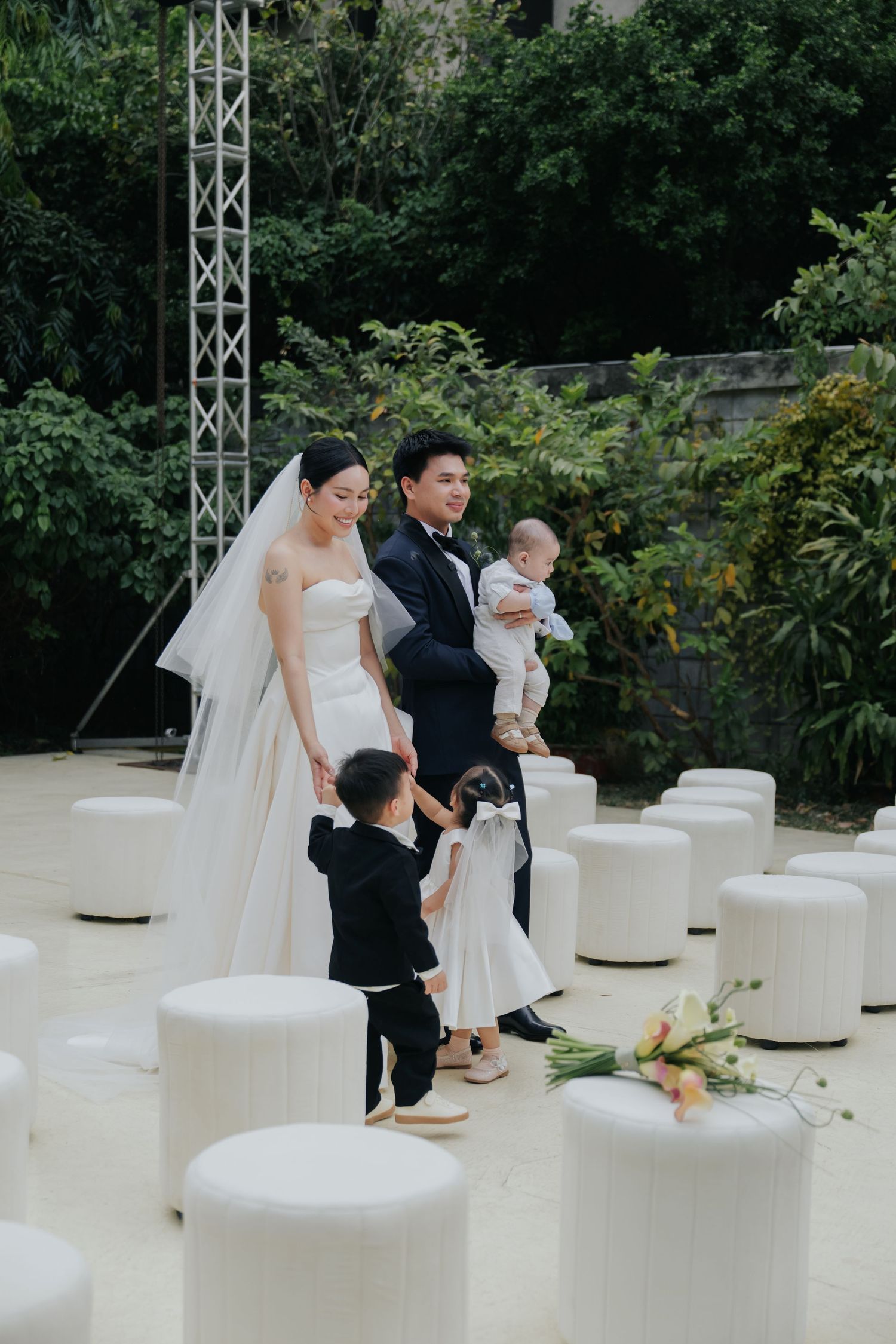 Bride and groom with children at outdoor wedding ceremony, white ottoman seating, lush greenery backdrop.