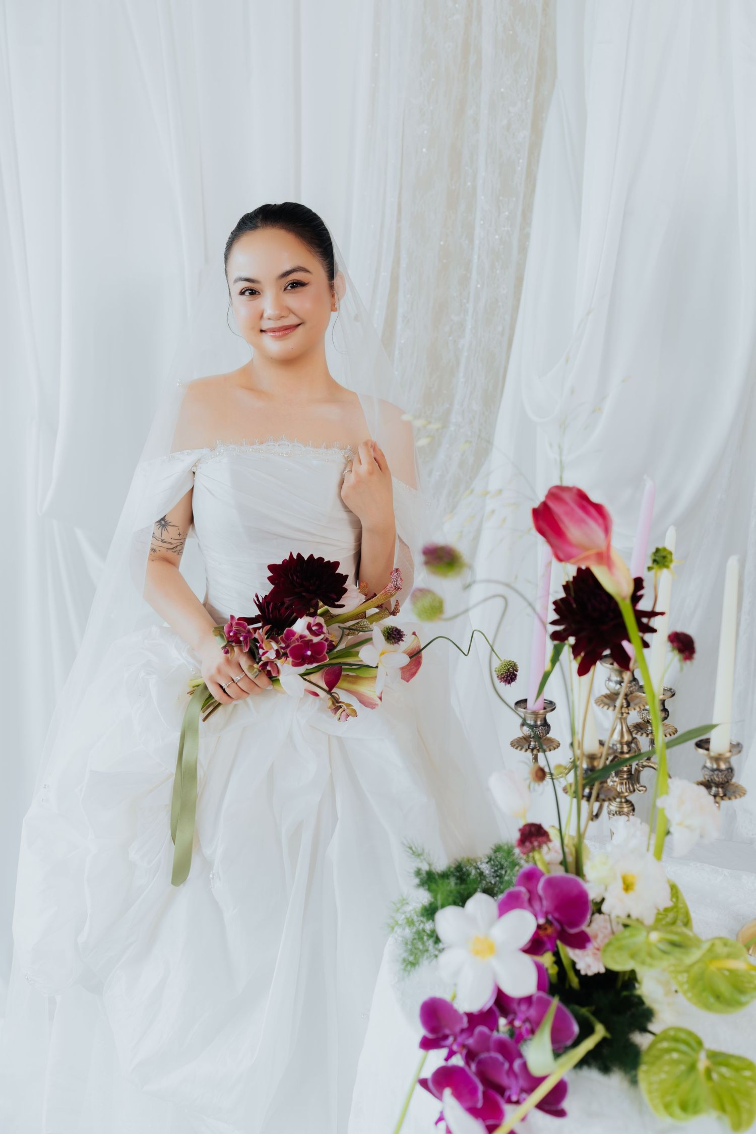 Smiling bride in white off-shoulder gown holding dark floral bouquet, standing near elegant flower arrangements against sheer curtains.