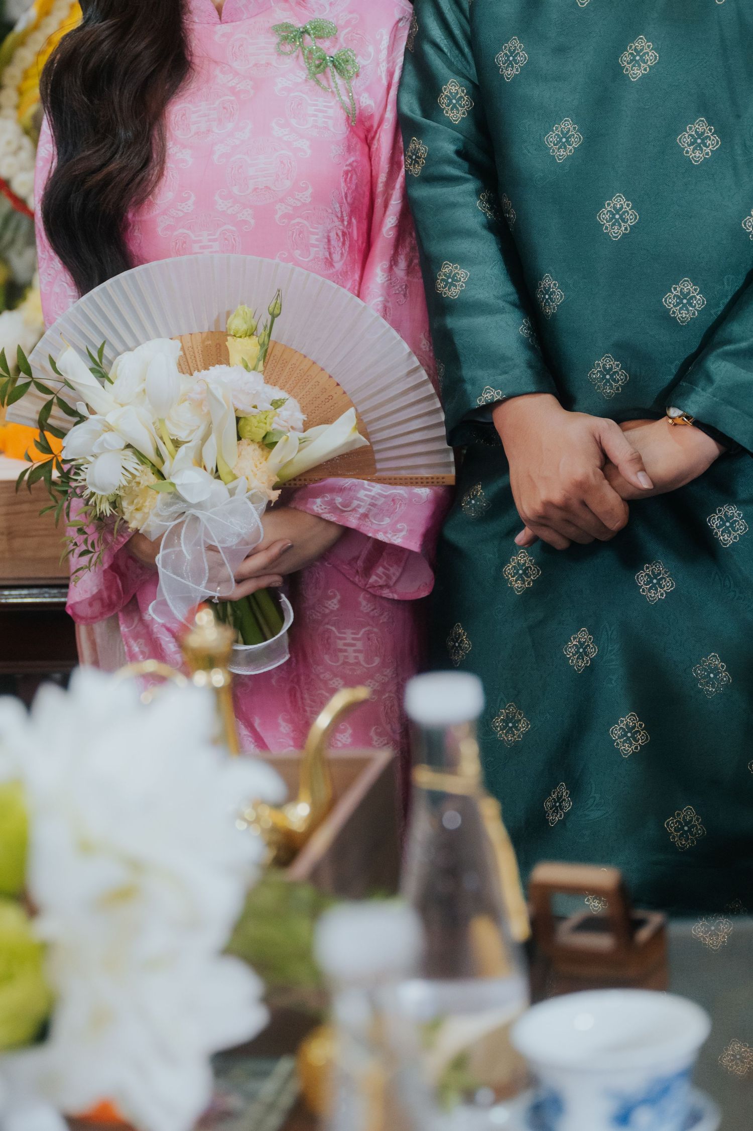 Couple in traditional Vietnamese ao dai attire, pink and teal brocade, holding white flower bouquet and fan at ceremony.