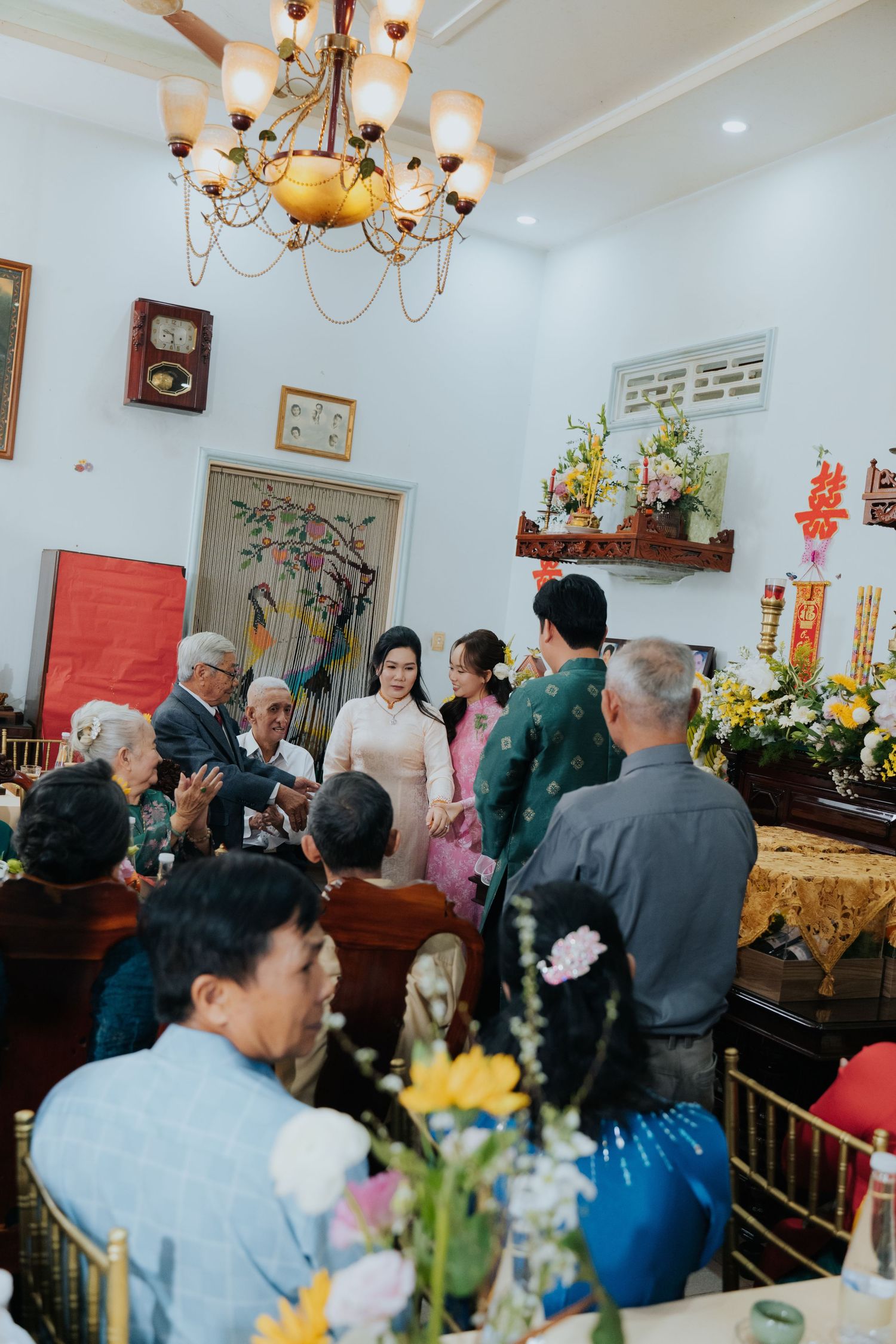Vietnamese wedding ceremony with guests in traditional ao dai attire gathered in a decorated home with floral arrangements.