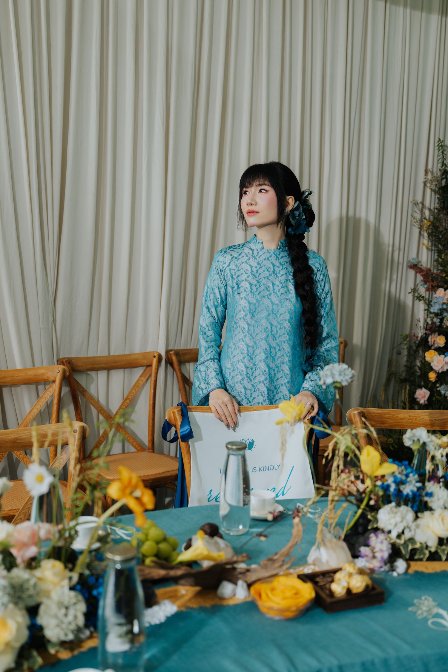 Young woman in light blue lace ao dai dress standing at a floral decorated table with colorful arrangements.