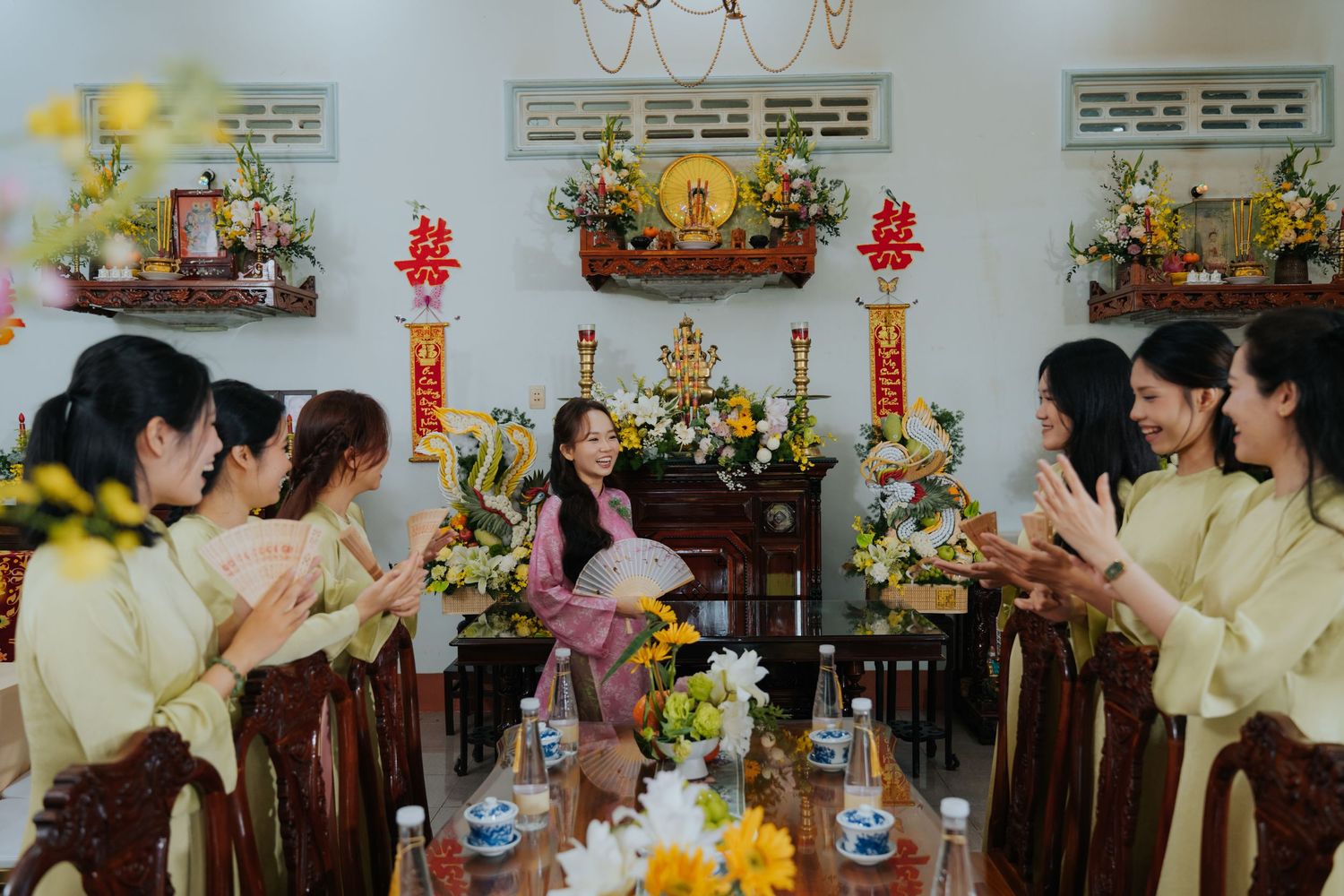 Young woman in pink ao dai celebrated by friends in yellow dresses at a traditional Vietnamese altar.