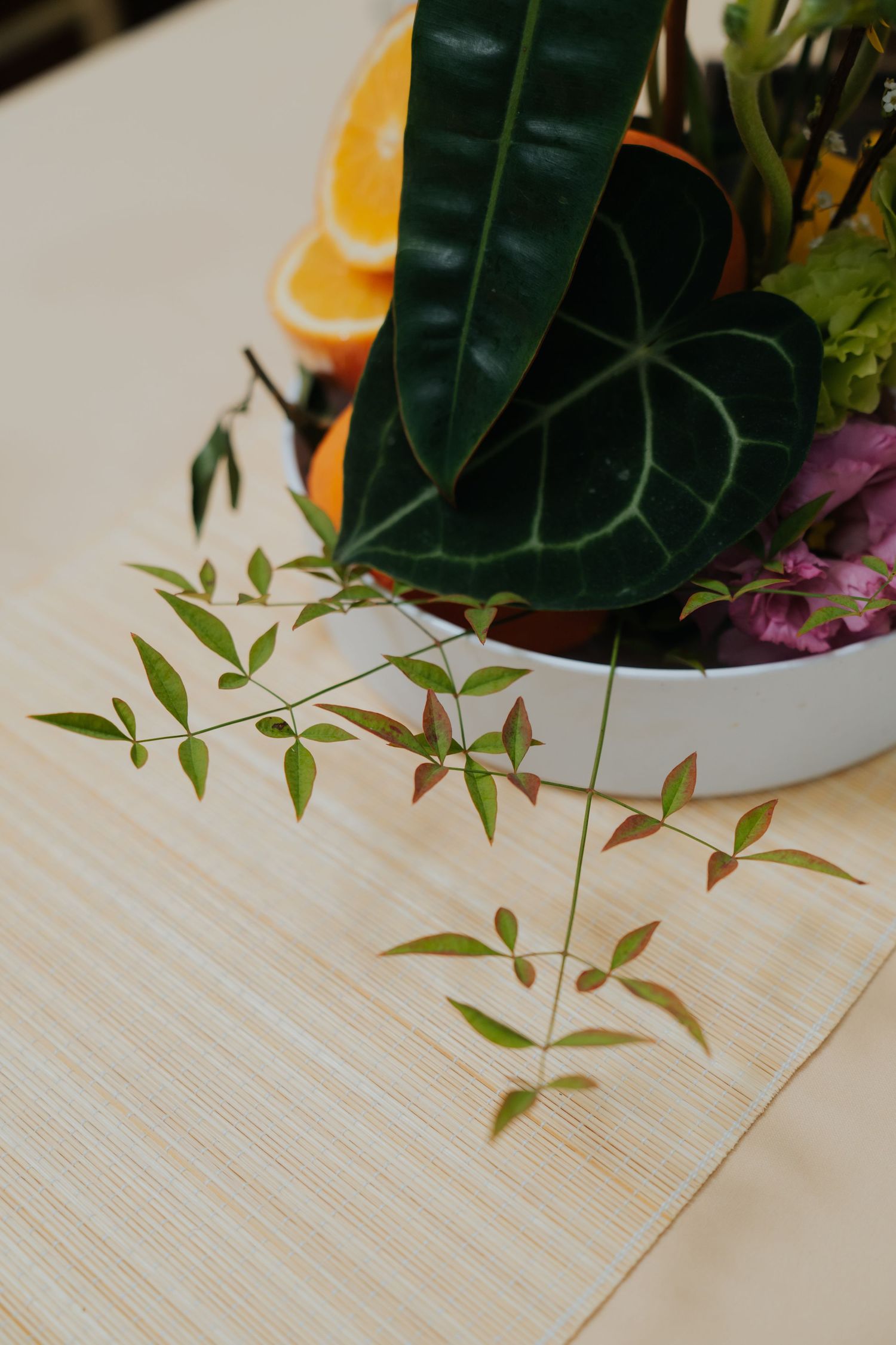 A floral arrangement with tropical leaves, orange slices, and pink flowers in a white bowl on a light wooden table.