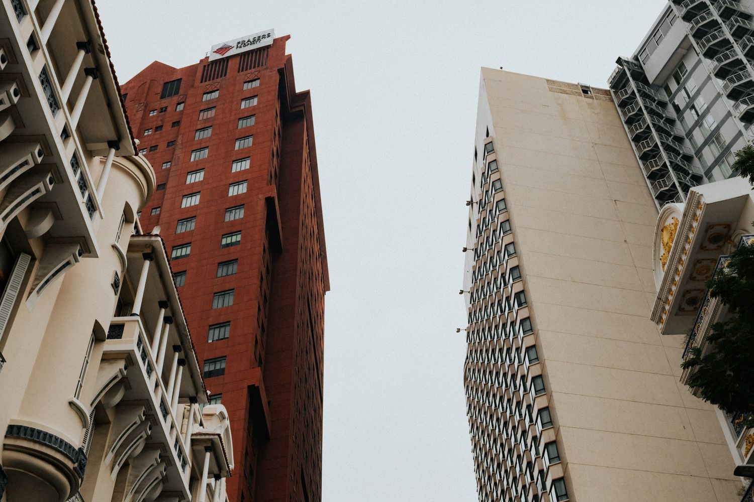 Low-angle view of tall urban buildings including a red brick skyscraper against a gray sky in a dense city.