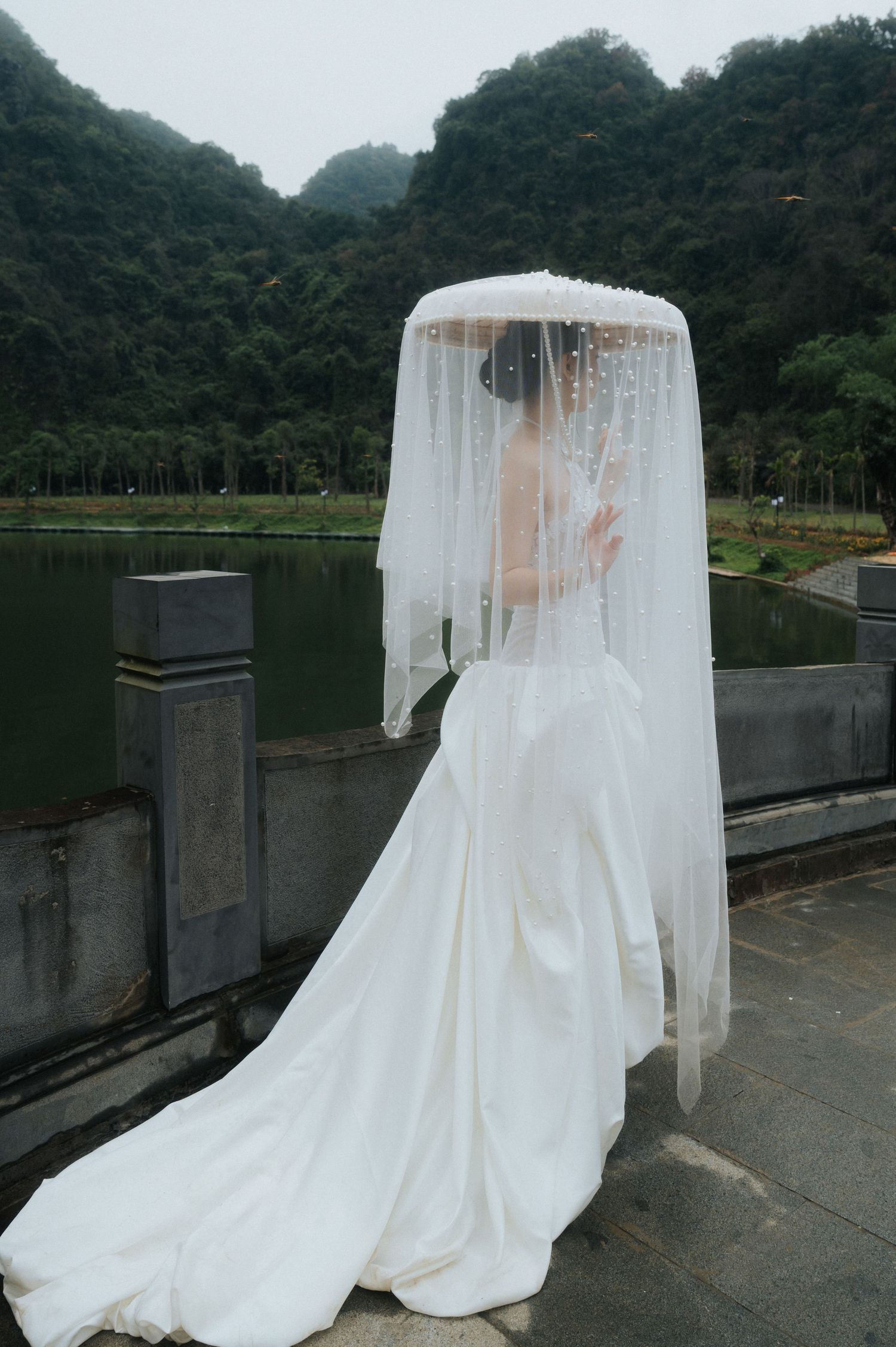 Bride in white wedding gown with pearl-adorned veil poses by a scenic lake with lush green mountains in background.