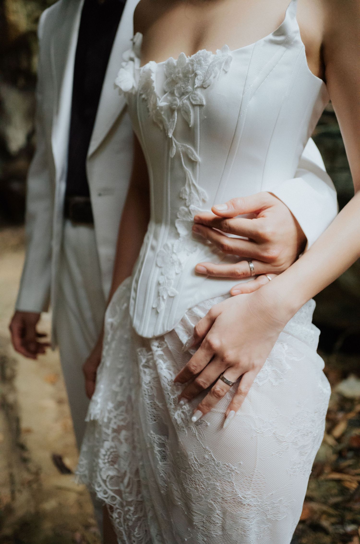 Bride in white corset lace wedding gown with groom's hands around her waist, showing wedding ring detail.