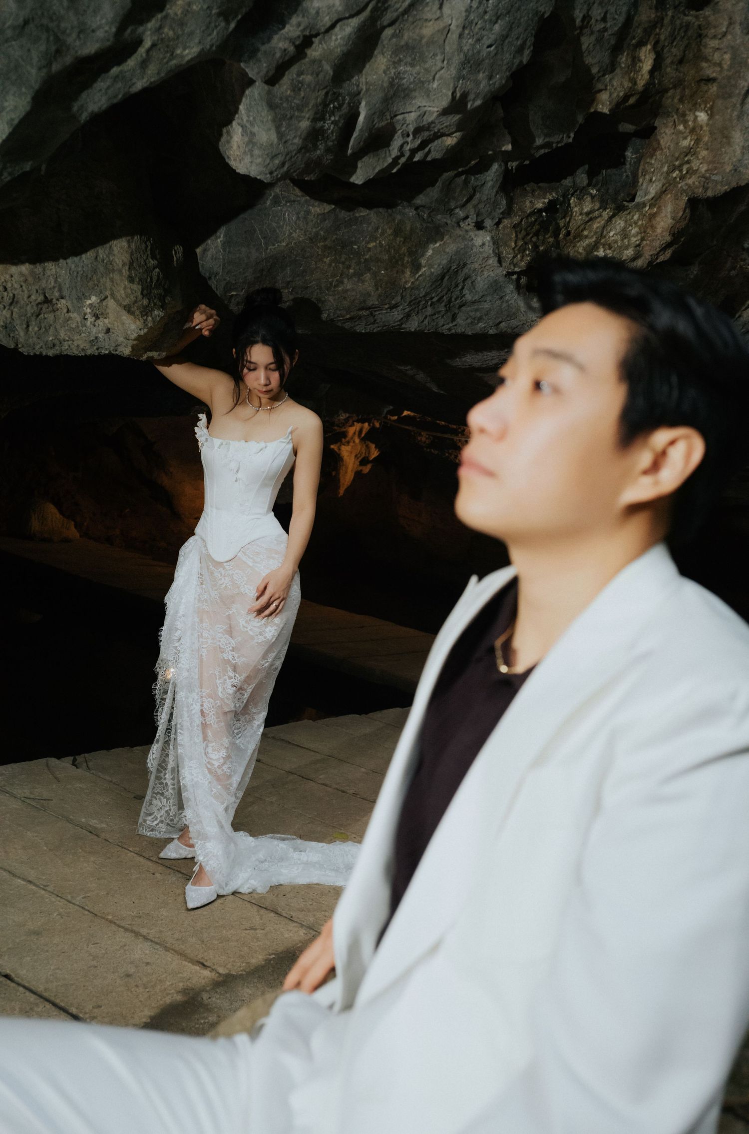 Groom in white suit sits in foreground as bride in lace wedding gown poses in rocky cave backdrop.