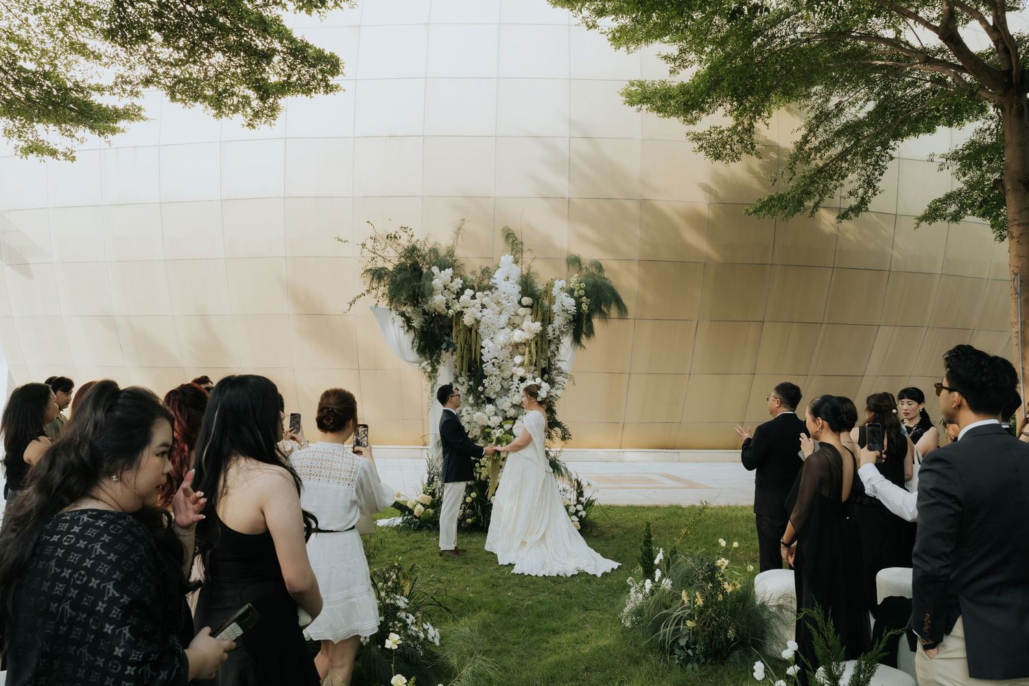Outdoor wedding ceremony with guests watching couple exchange vows beneath a floral arch on a manicured lawn.