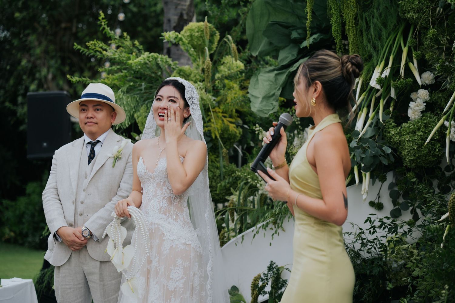 Bride laughing joyfully during outdoor wedding ceremony with groom and officiant amid lush tropical greenery.