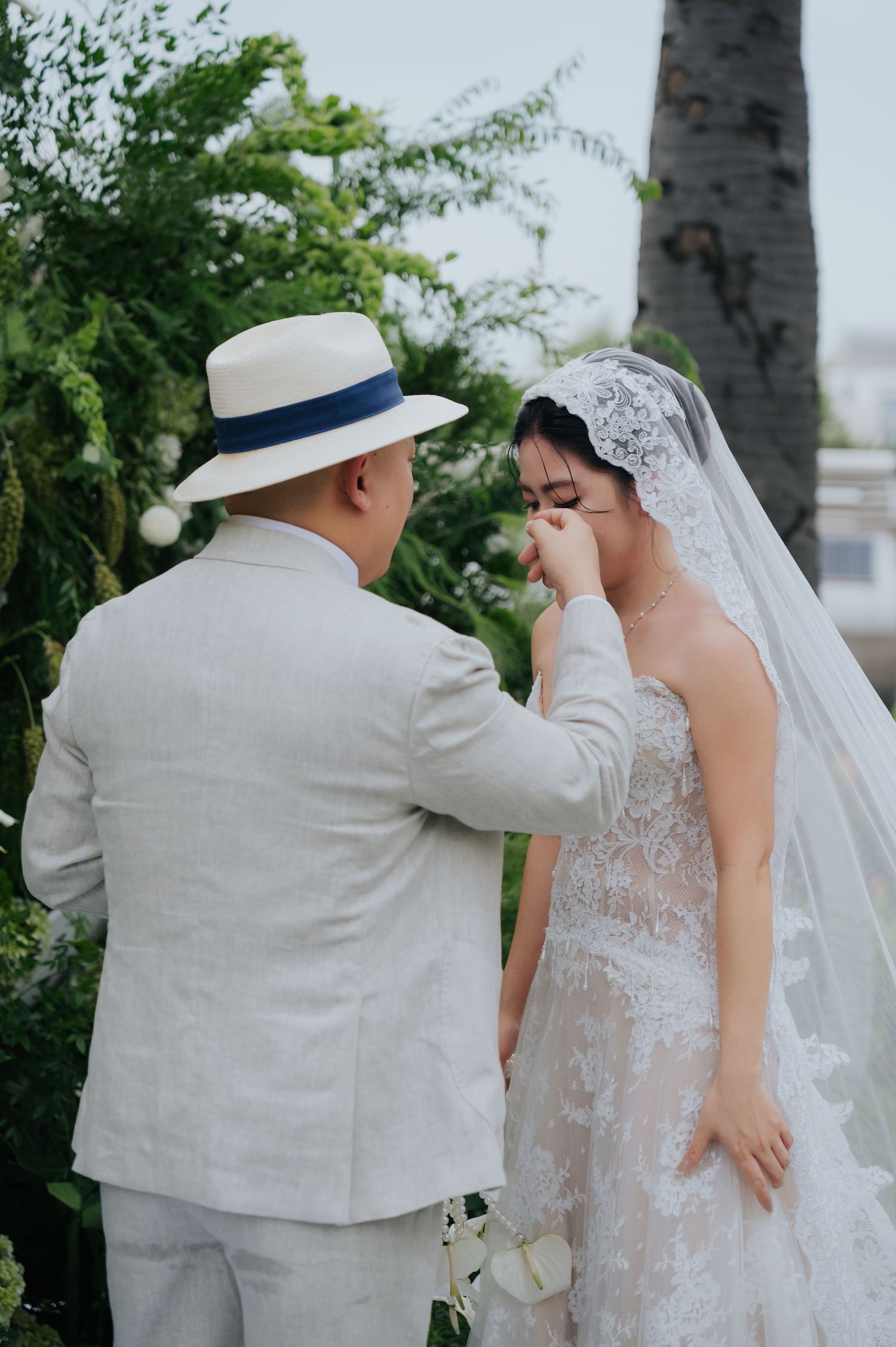 Groom in linen suit and panama hat gently wipes tears from emotional bride in lace wedding dress during outdoor ceremony.