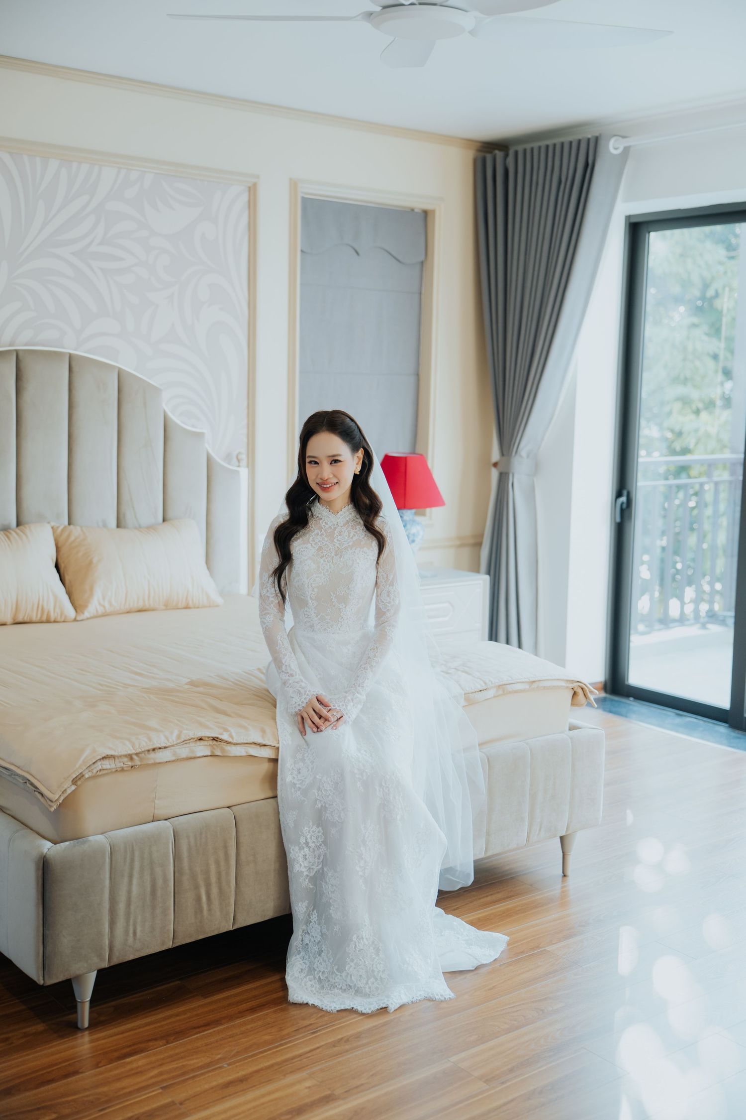 A smiling bride in a white lace long-sleeve wedding dress stands elegantly in a bright, modern bedroom.