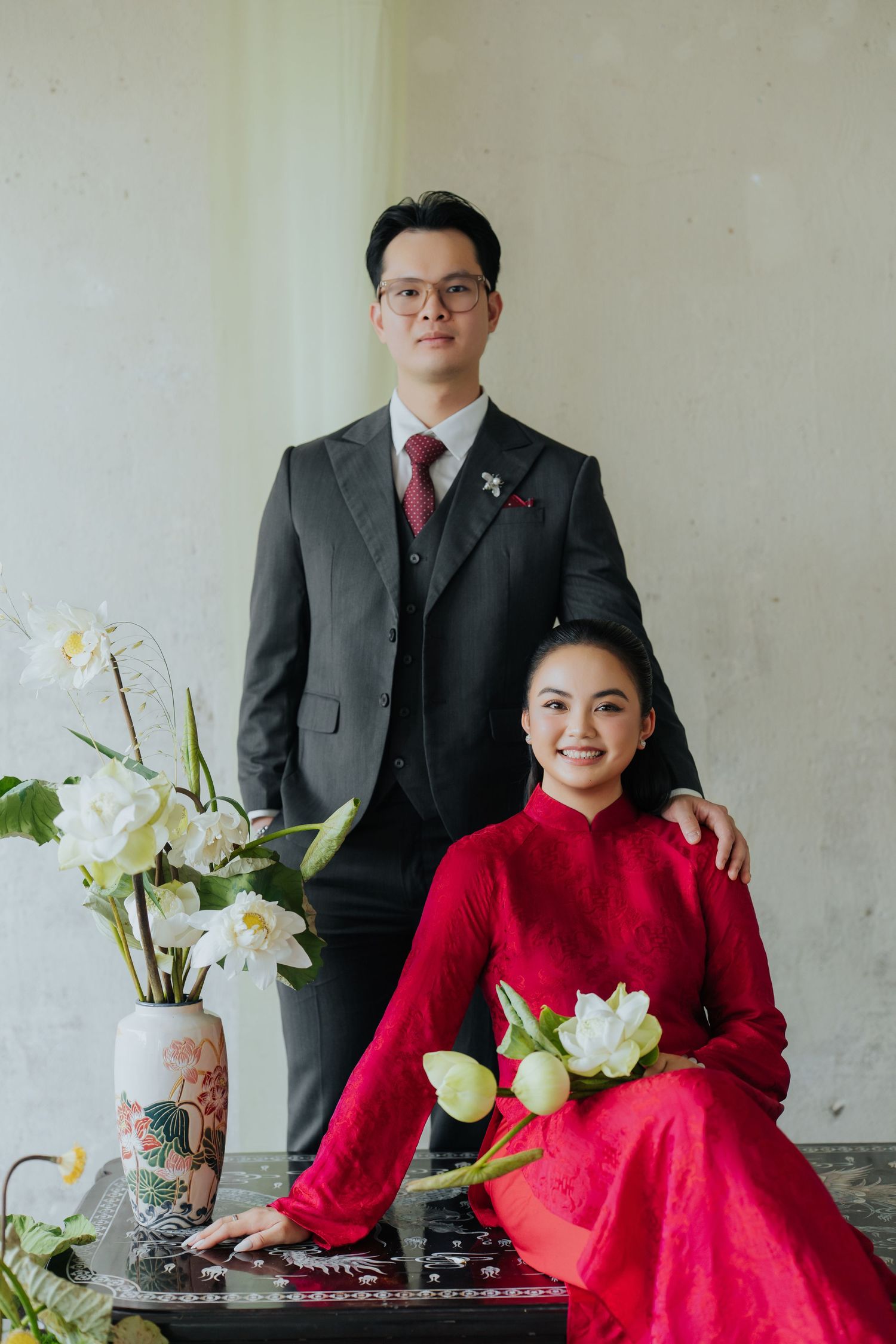 Vietnamese couple in traditional ao dai and suit posing with white lotus flowers for a formal portrait.