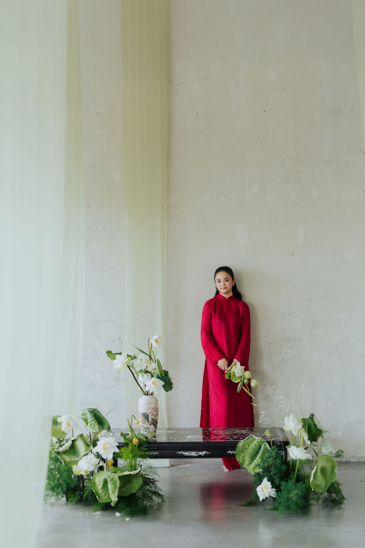 Woman in red dress stands on platform surrounded by white flowers and greenery in minimalist space.