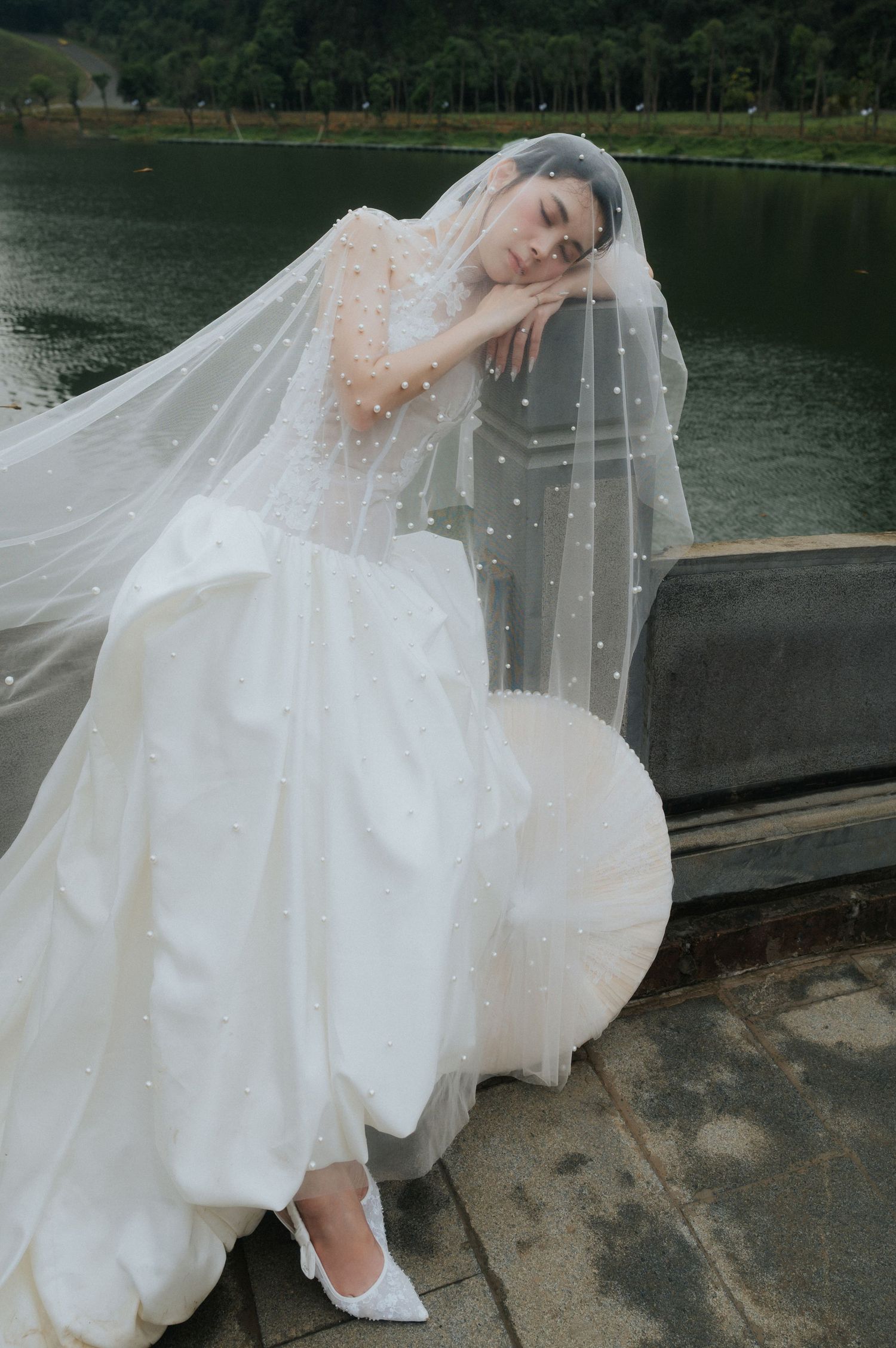Bride in a white ball gown with pearl-embellished veil poses gracefully by a serene lakeside.