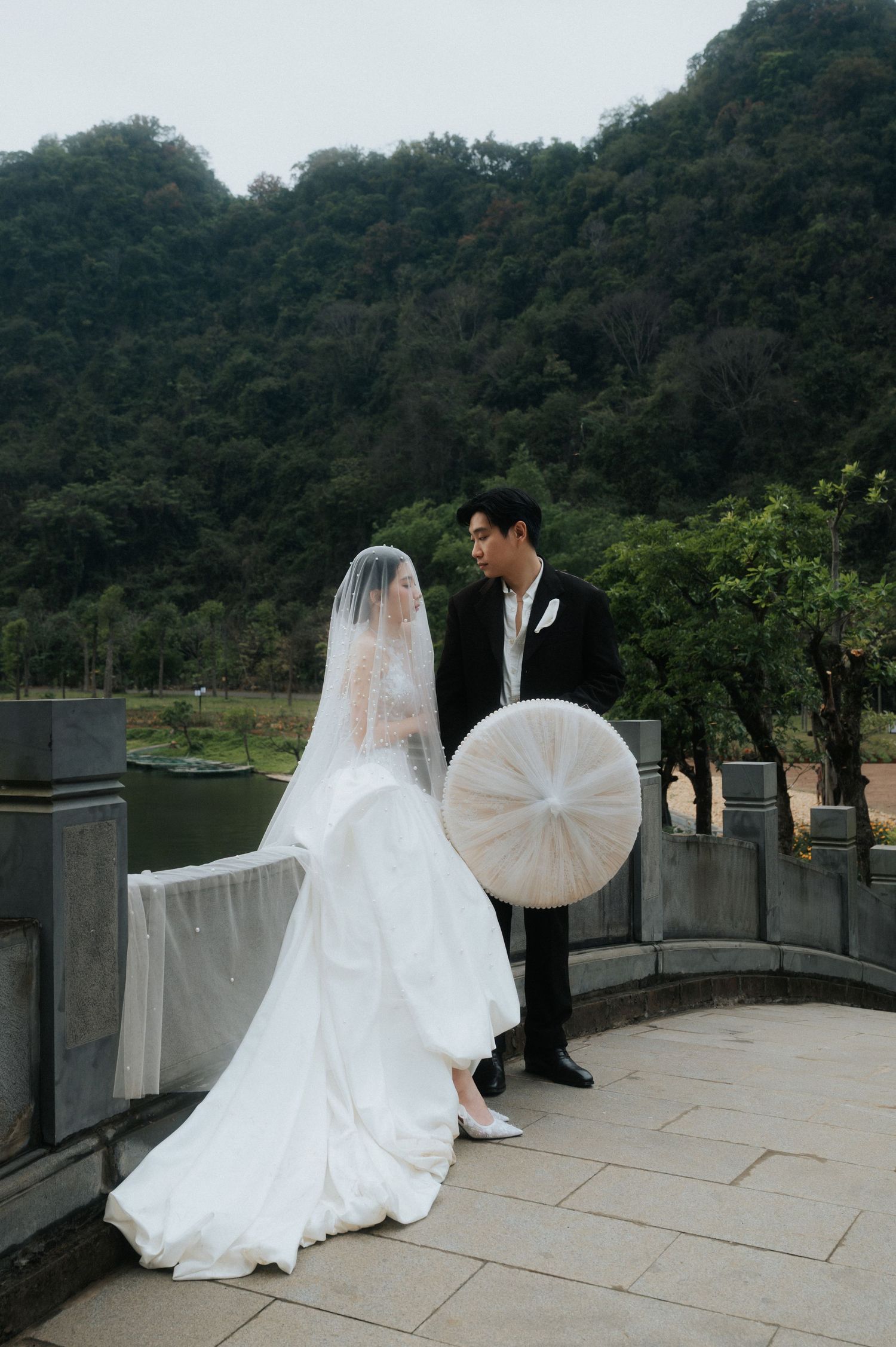 A bride in a white gown and veil stands with a groom in a black suit on a scenic outdoor terrace with lush green hills.