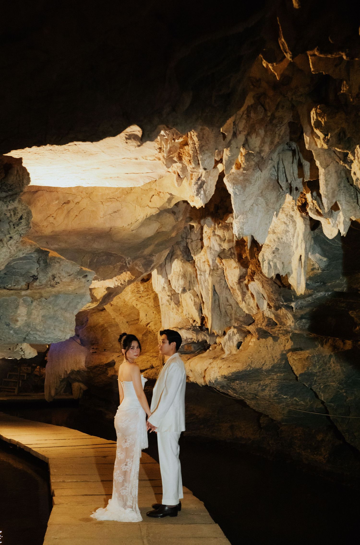 A couple in wedding attire holds hands inside a dramatic illuminated limestone cave with stunning rock formations.