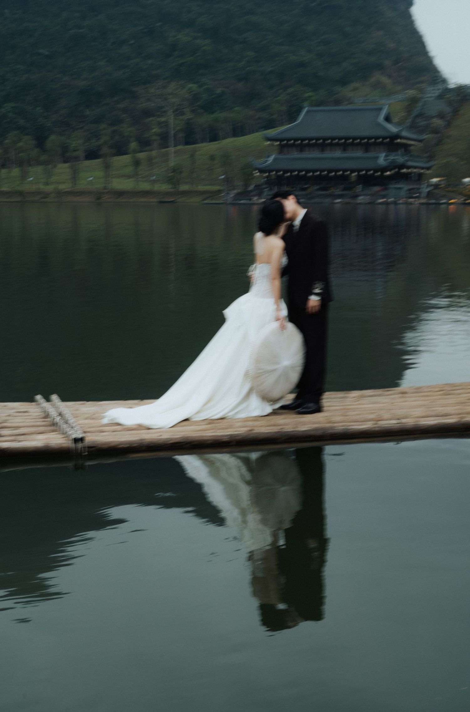 A bride and groom share a romantic kiss on a wooden dock over a calm lake with an Asian temple in the background.