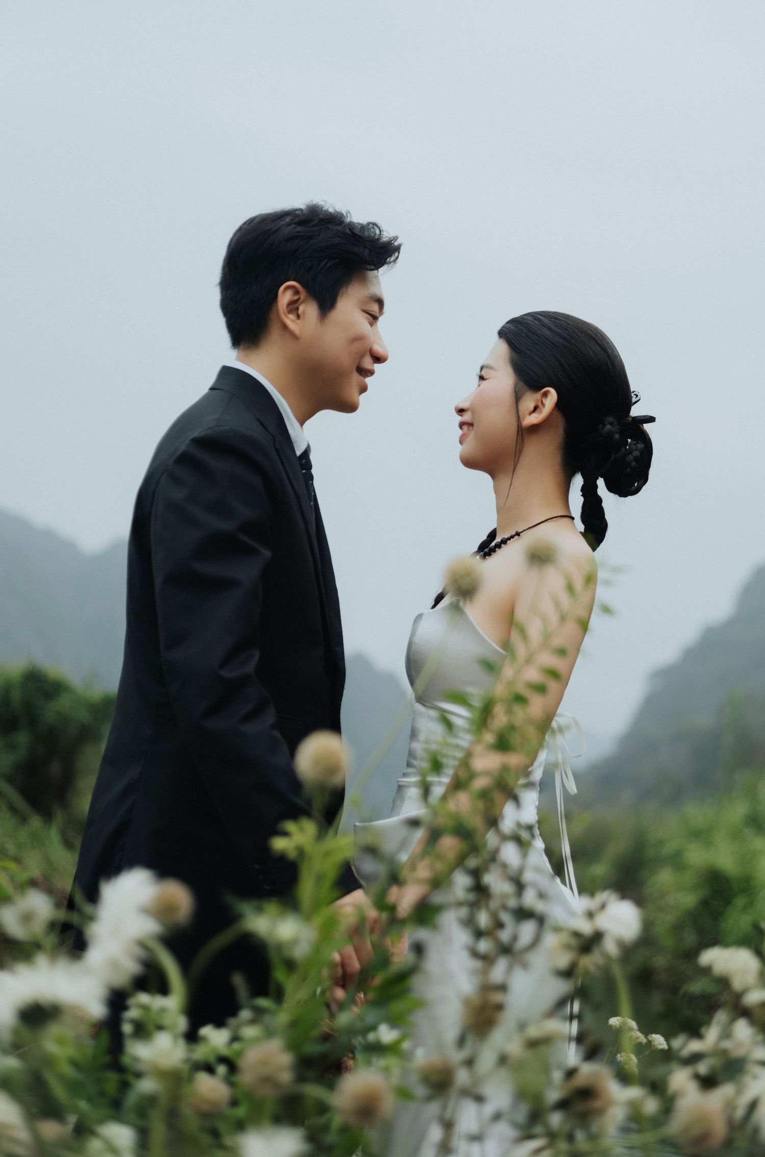 A couple in wedding attire smiles at each other surrounded by wildflowers with misty mountains in the background.