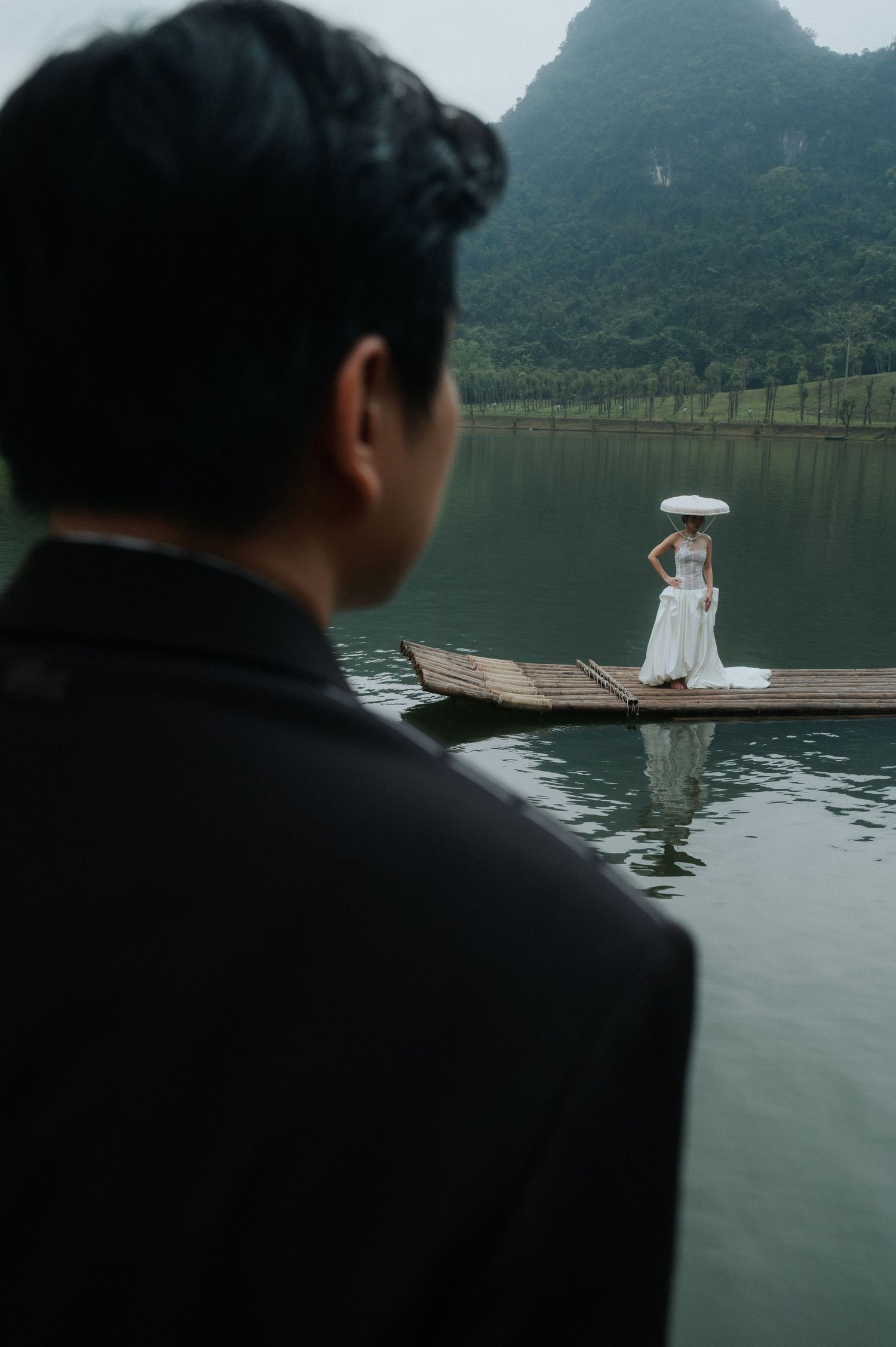 A groom in a black suit watches his bride in a white dress standing on a wooden boat on a serene lake in Vietnam.