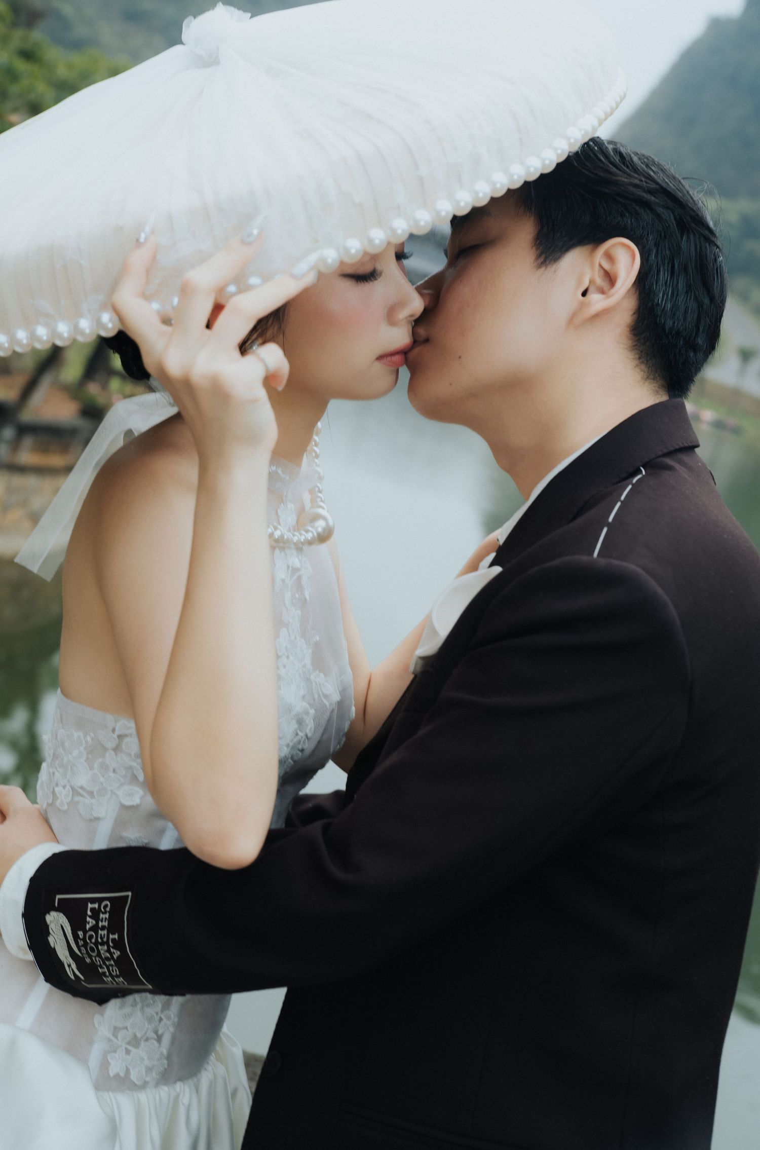 A couple shares a romantic kiss under a white pearl-trimmed umbrella in wedding attire outdoors.