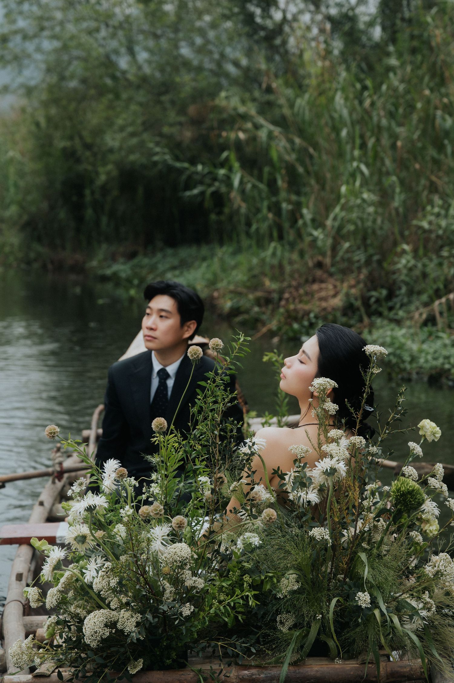 Couple in a flower-filled boat on a serene river, surrounded by lush greenery, in elegant wedding attire.