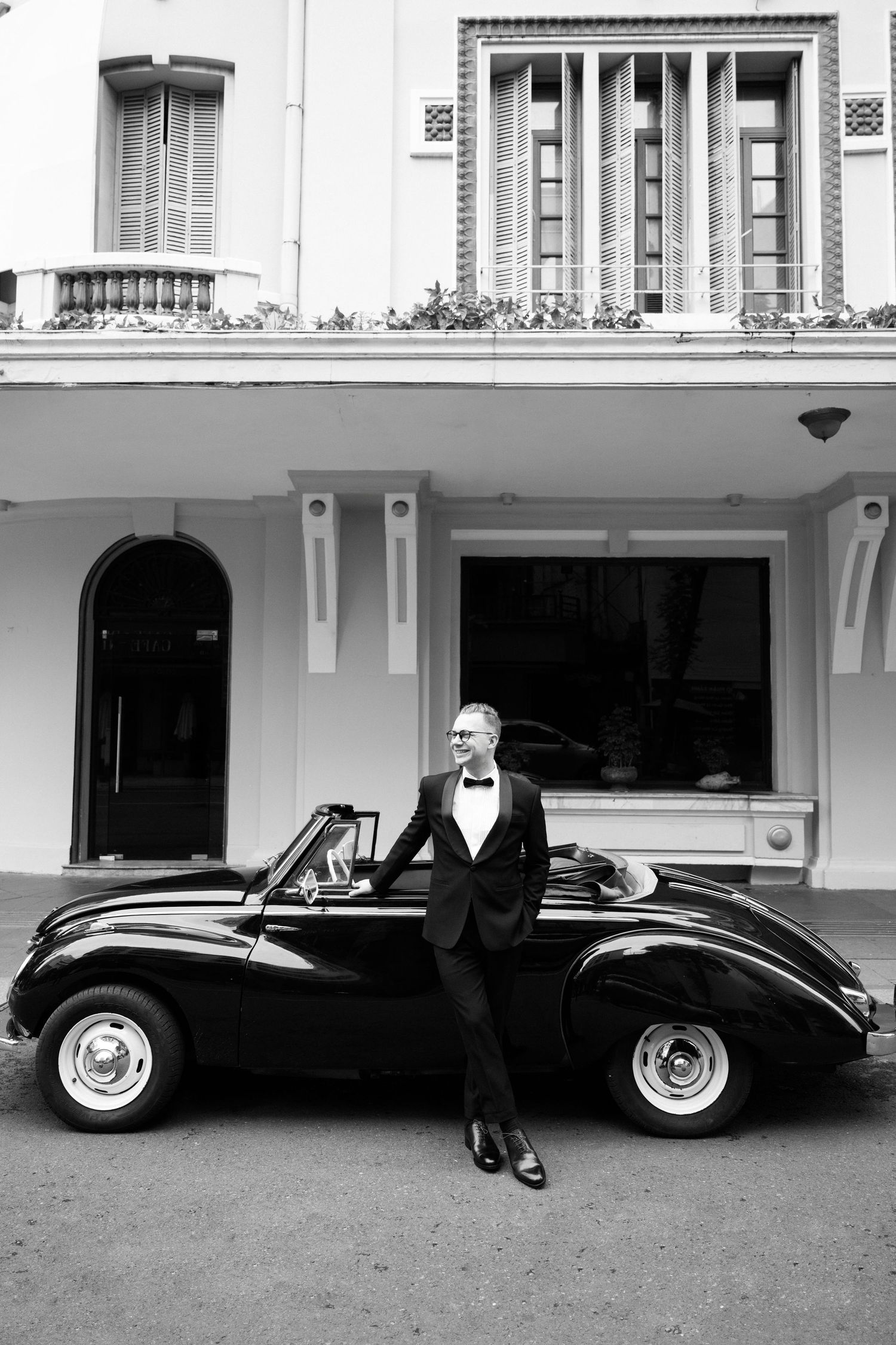A dapper man in a tuxedo leans against a vintage convertible car outside an elegant building in black and white.