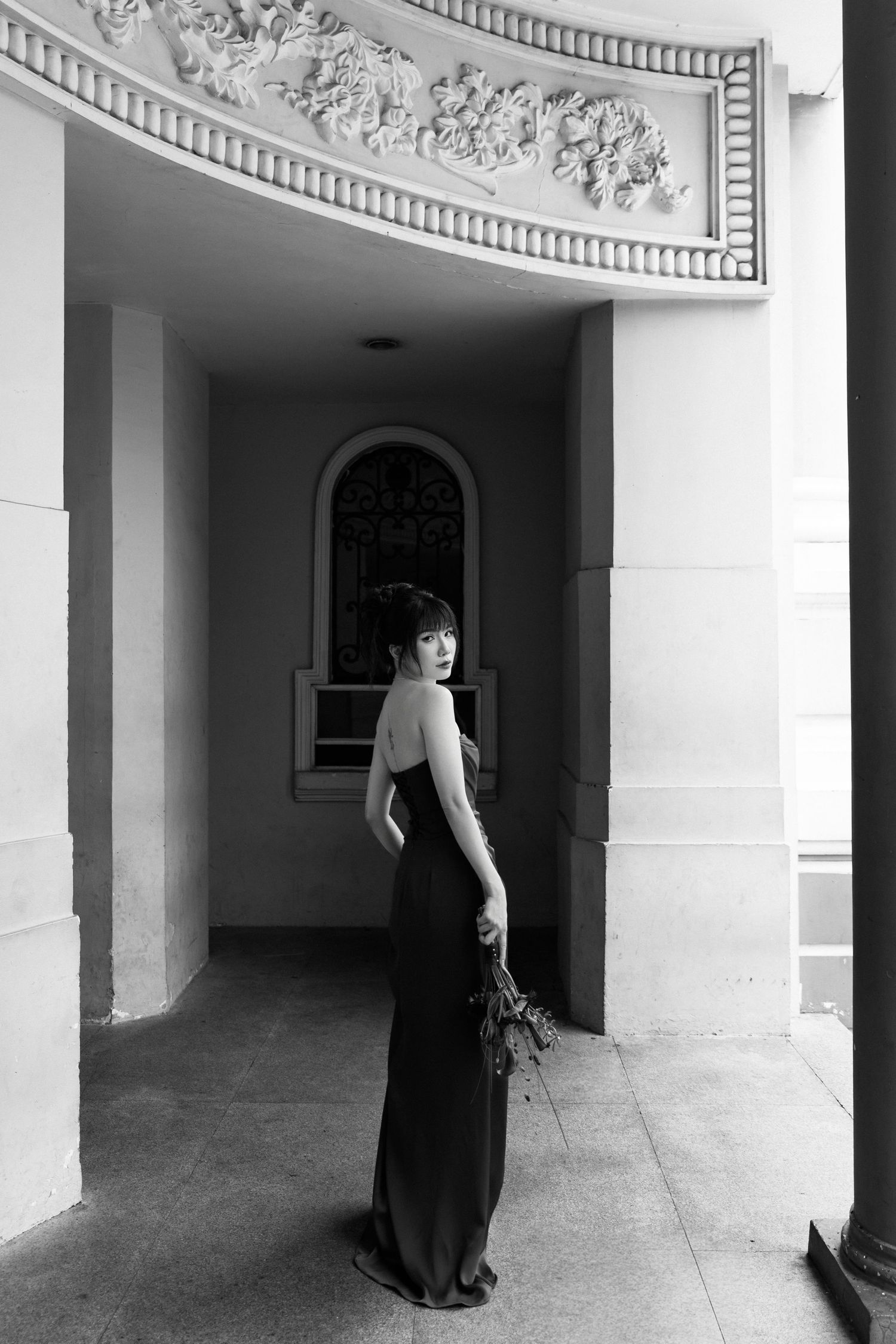 Elegant woman in black gown holding flowers poses in ornate architectural archway in black and white.