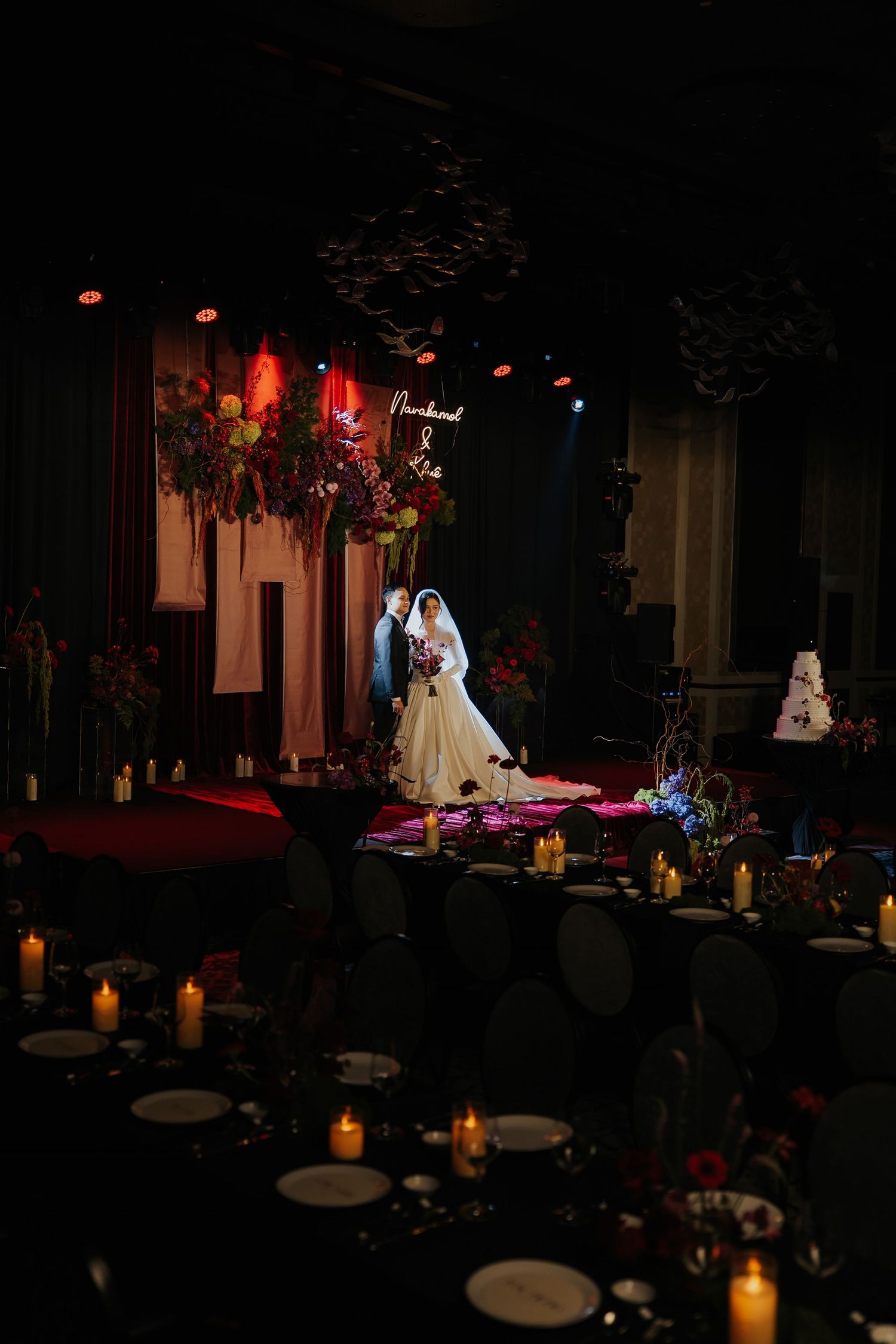 Bride and groom stand at a dramatic dark wedding reception with candlelit tables, floral backdrop, and elegant stage lighting.