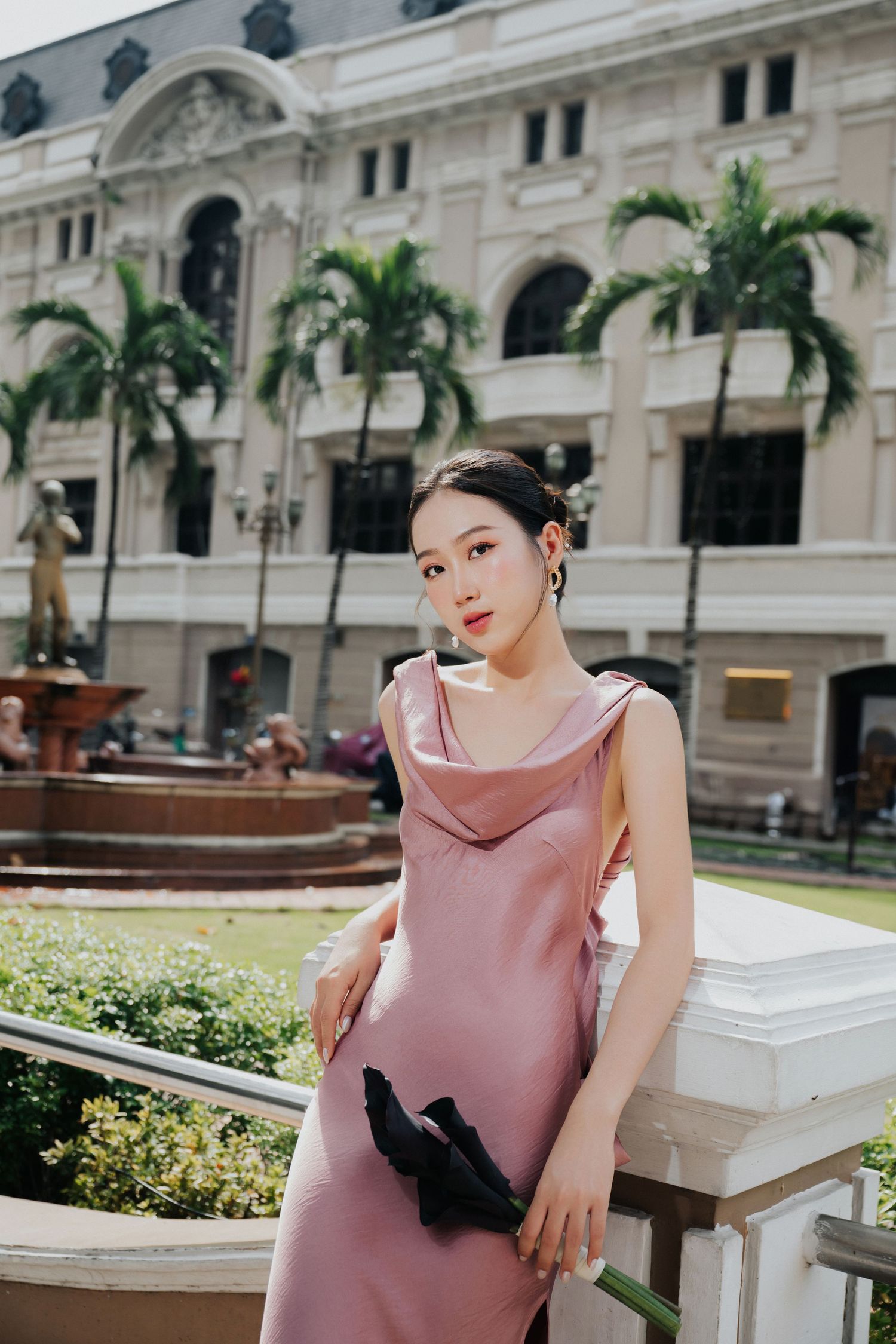 A young Asian woman in a pink satin dress poses gracefully by a white balustrade with palm trees and a fountain behind her.