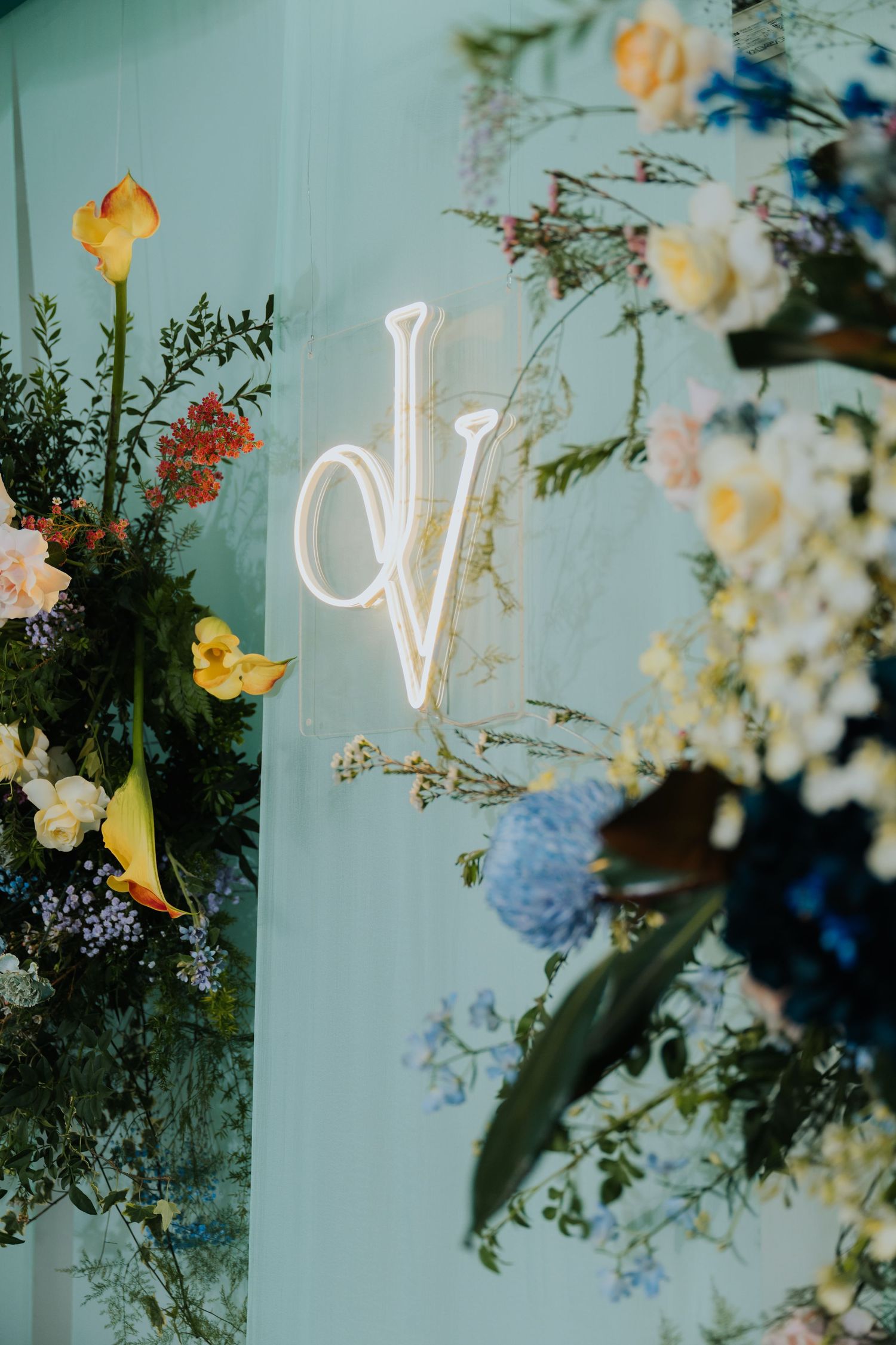 Neon monogram dV sign on mint wall surrounded by colorful floral arrangement with blue hydrangeas and yellow blooms.