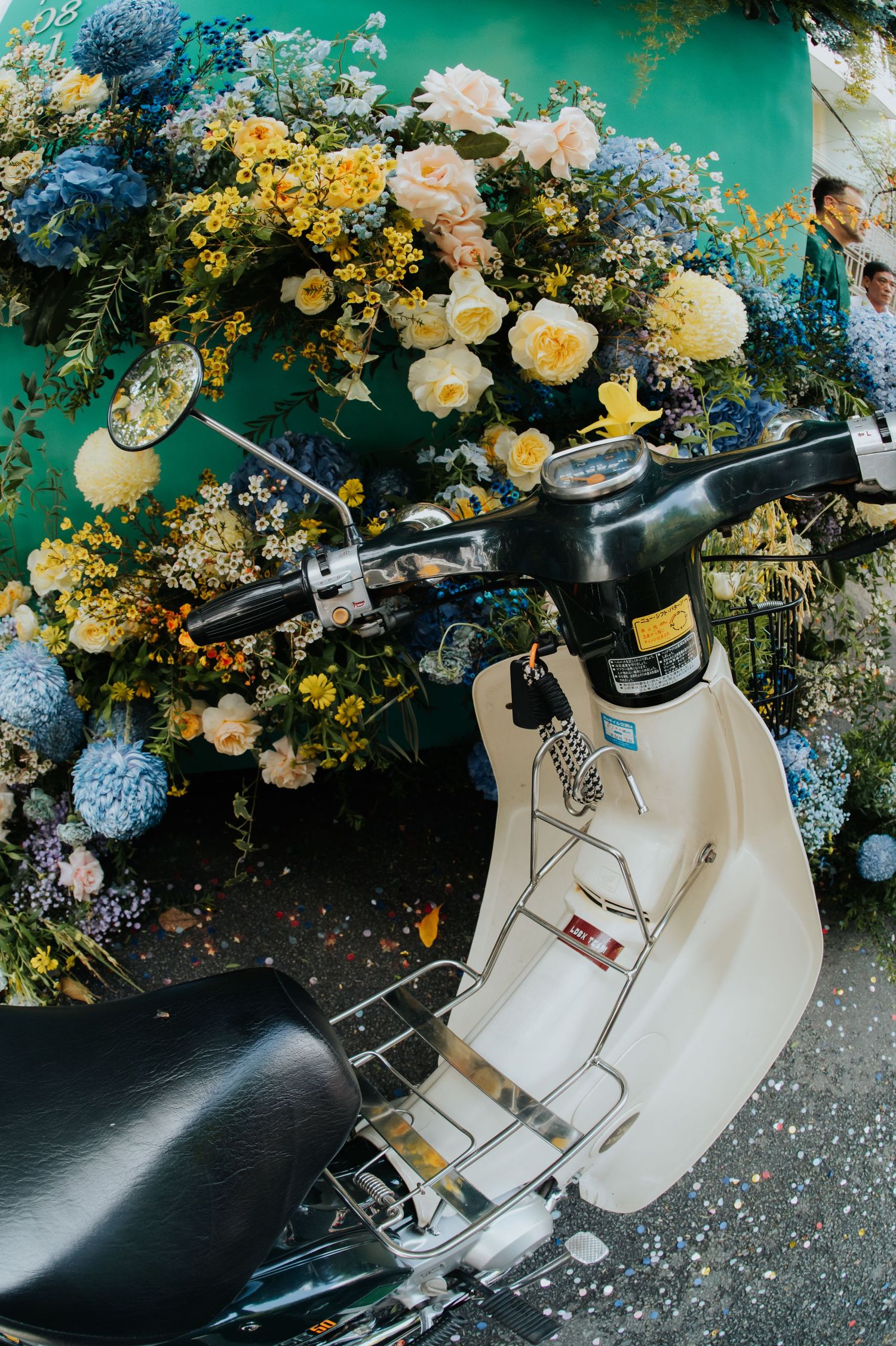 White vintage scooter displayed against a vibrant floral backdrop with yellow, blue, and white flowers.