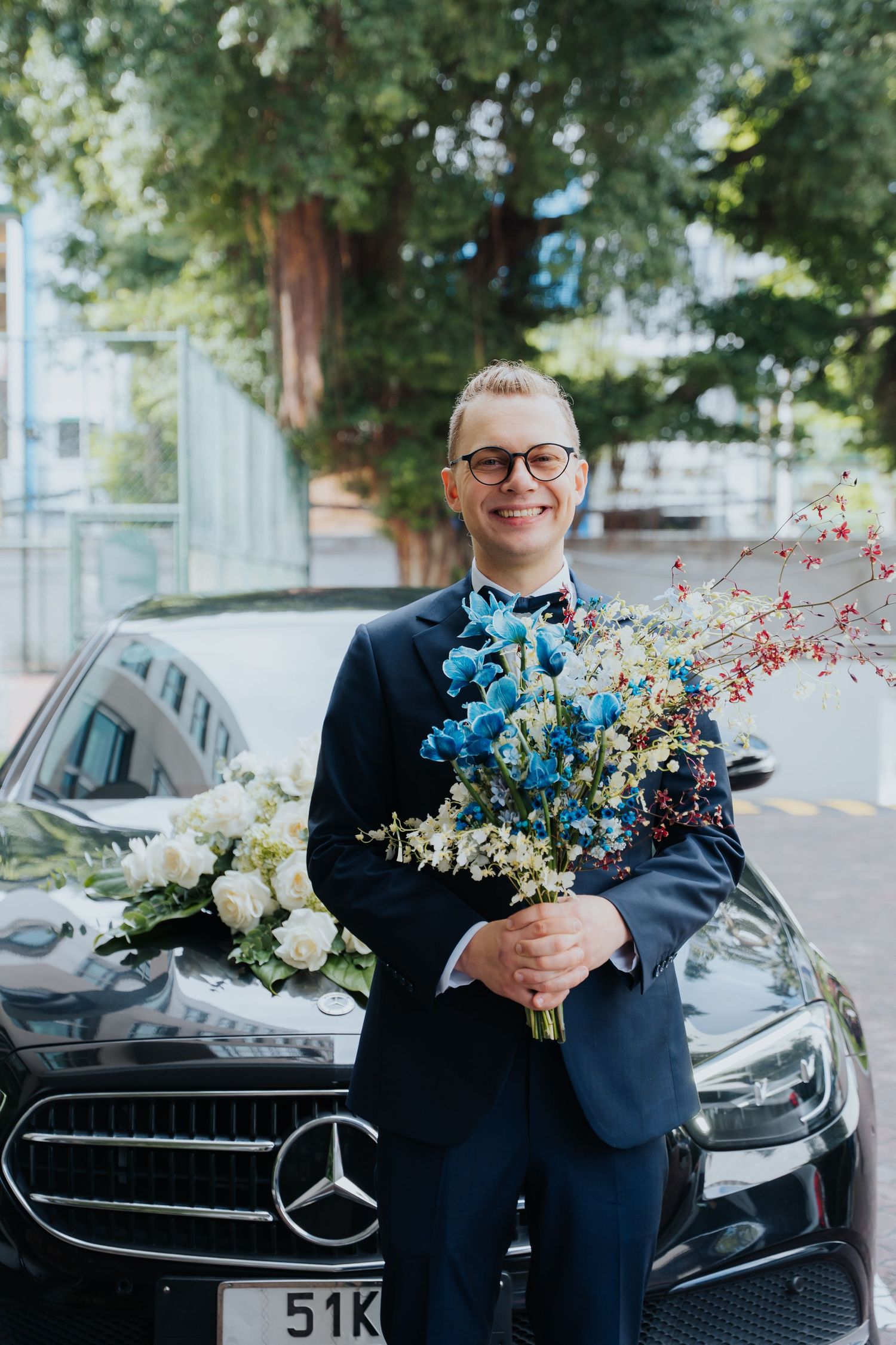 Smiling groom in navy suit holding blue and white wedding bouquet standing beside black Mercedes-Benz car.