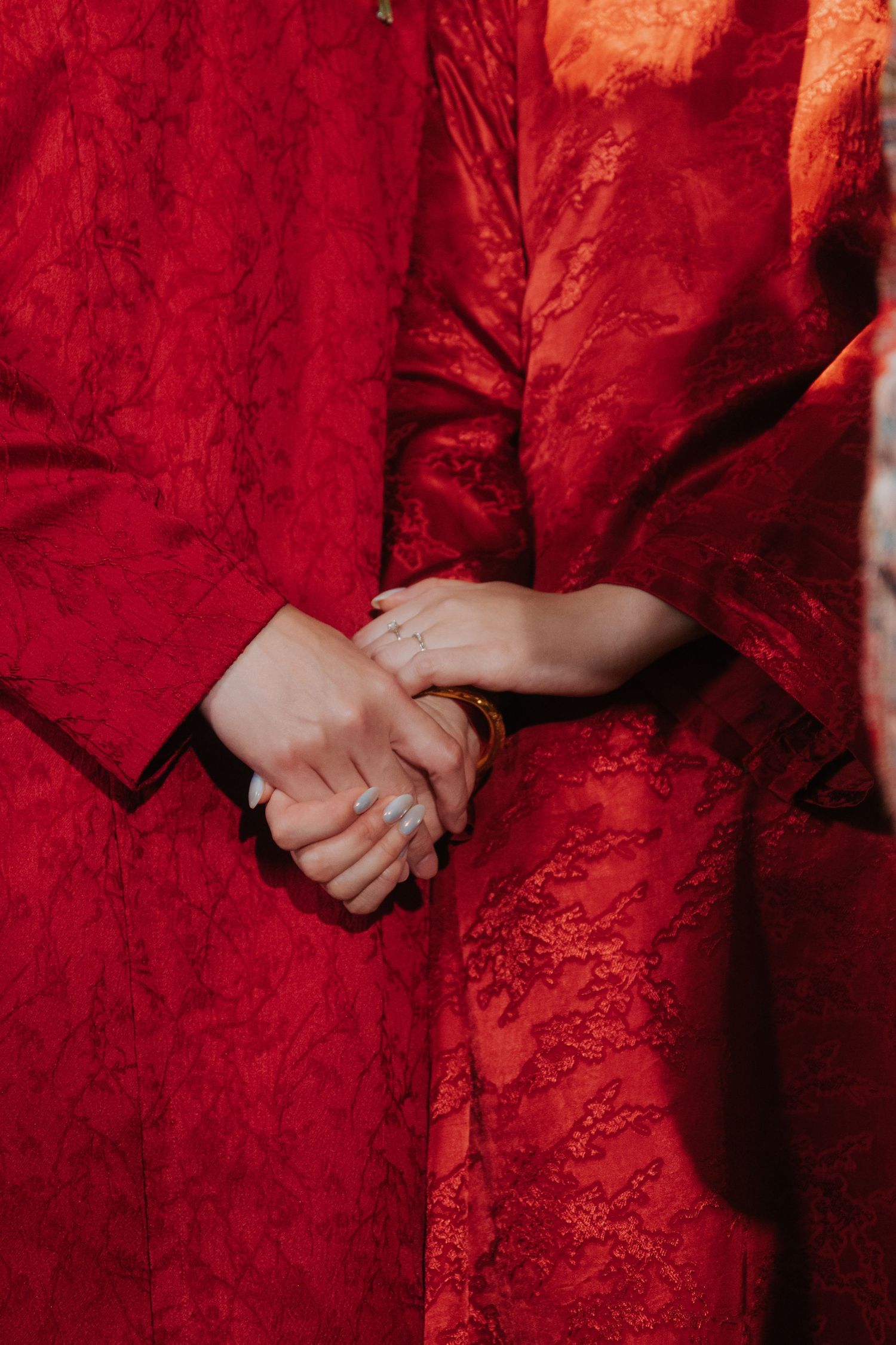 Two people in red lace traditional Chinese wedding attire holding hands, showcasing a gold bracelet and delicate ring.