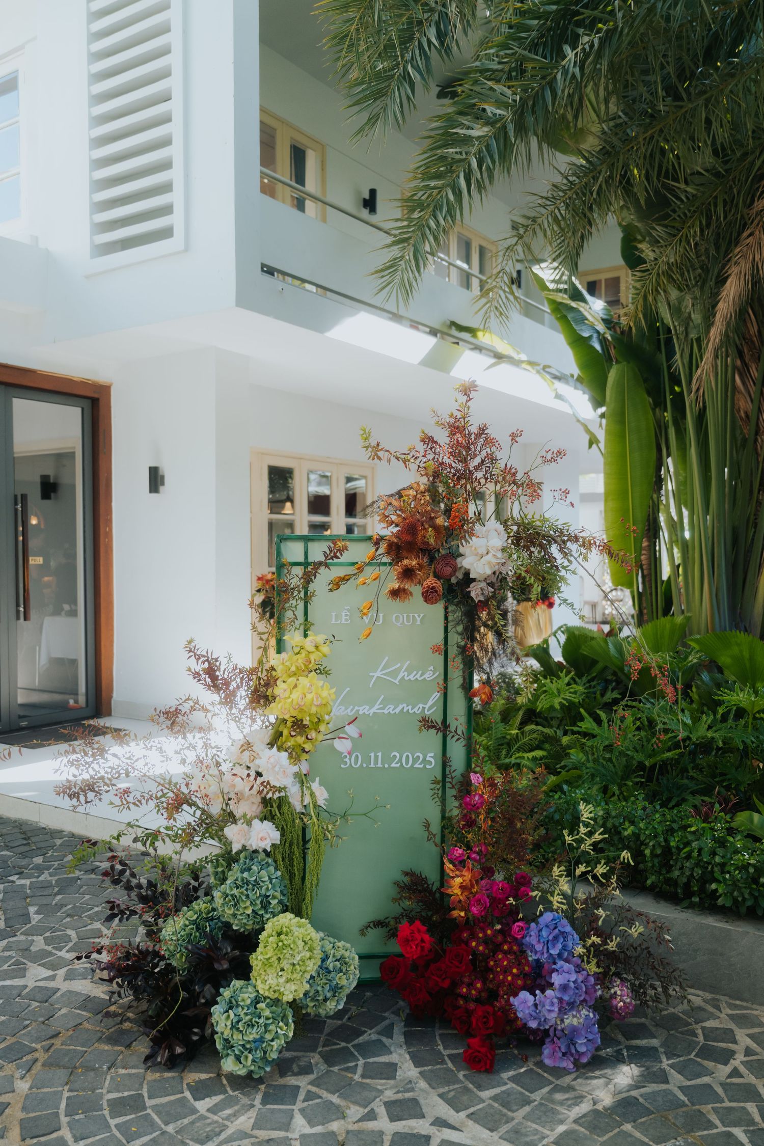 Elegant wedding welcome sign on green board surrounded by colorful floral arrangements outside a white tropical venue.