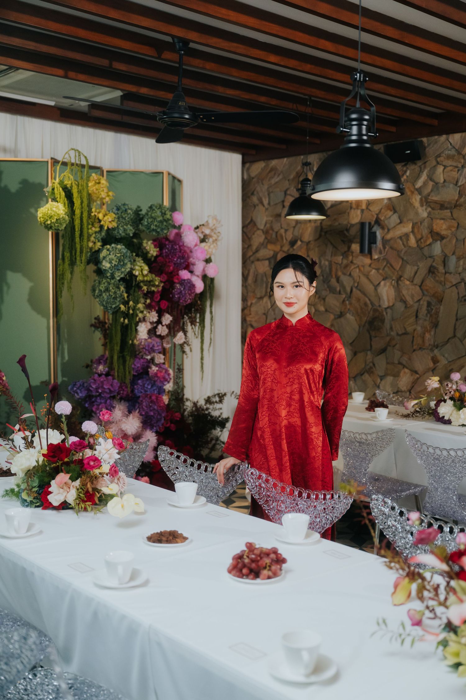 Woman in red traditional Vietnamese ao dai dress standing at decorated table with colorful floral arrangements.