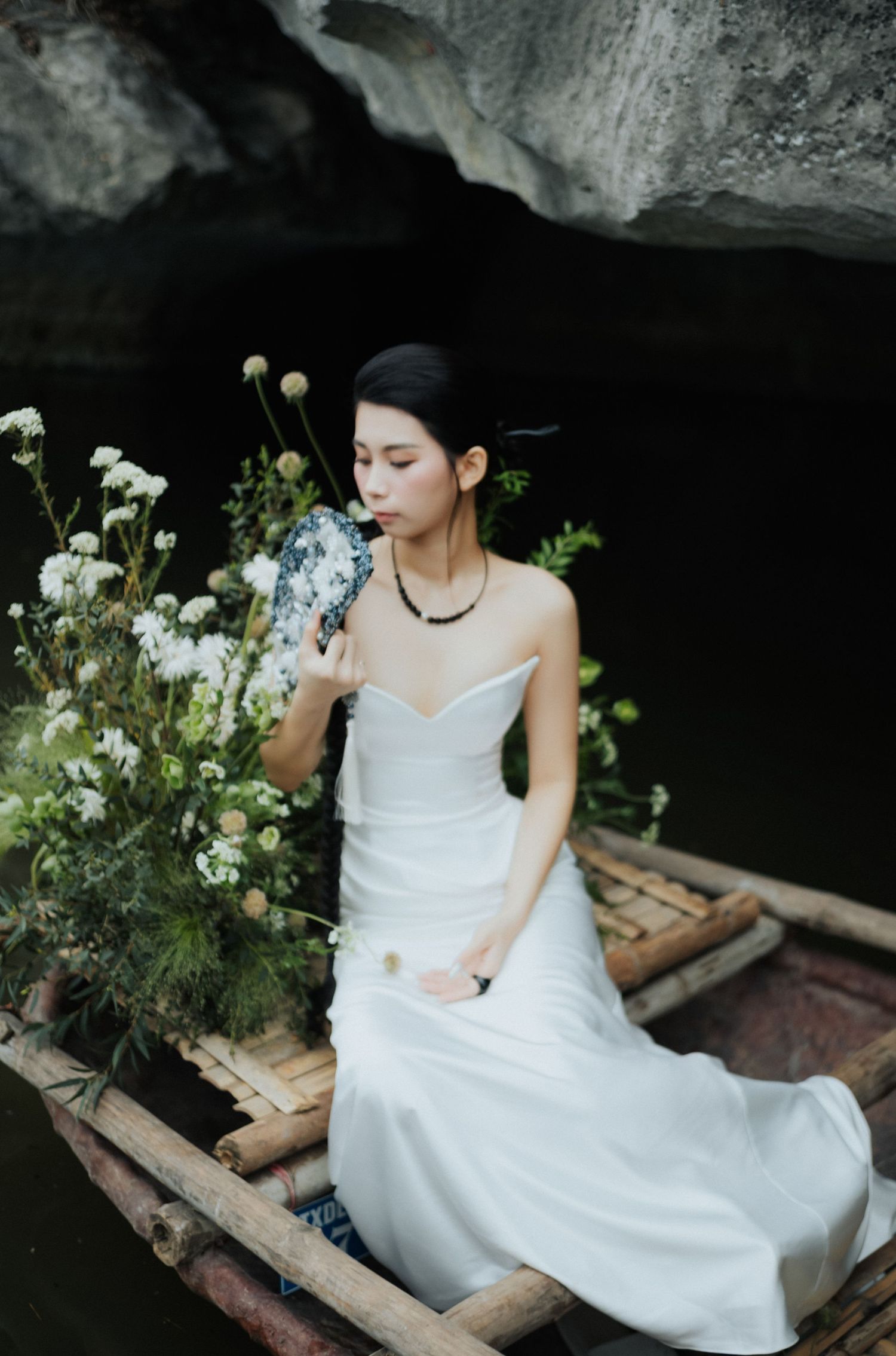 A woman in a white strapless gown sits in a bamboo boat, smelling white wildflowers near a rocky cave.