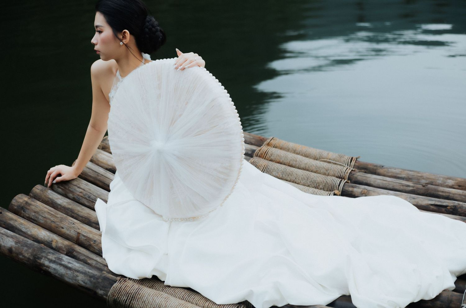 Bride in white gown holding a white parasol sits gracefully on a bamboo raft by calm dark water.