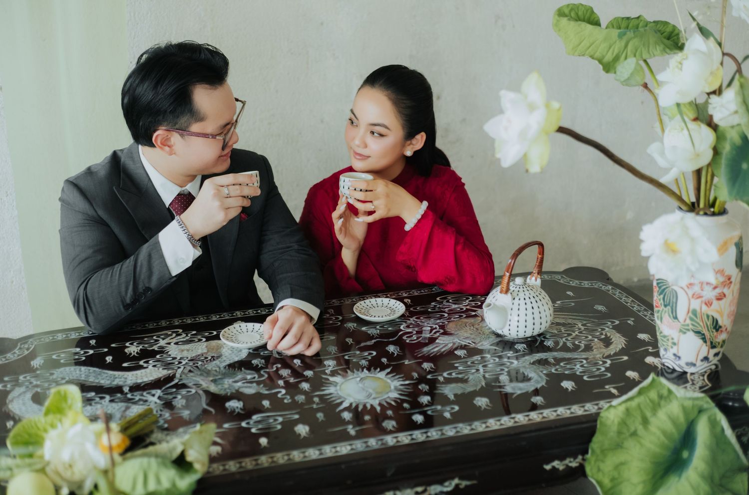 Couple sharing tea at ornate dark wooden table, woman in red dress, man in suit, with lotus flowers nearby.