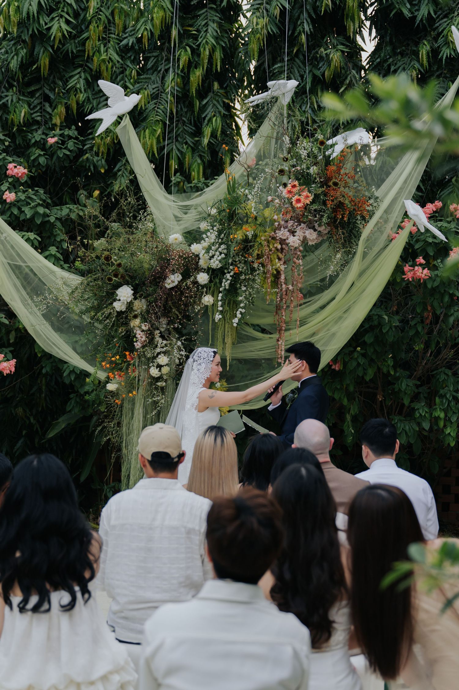 Outdoor wedding ceremony with bride and groom at floral arch, green draping, white doves, and lush garden backdrop.