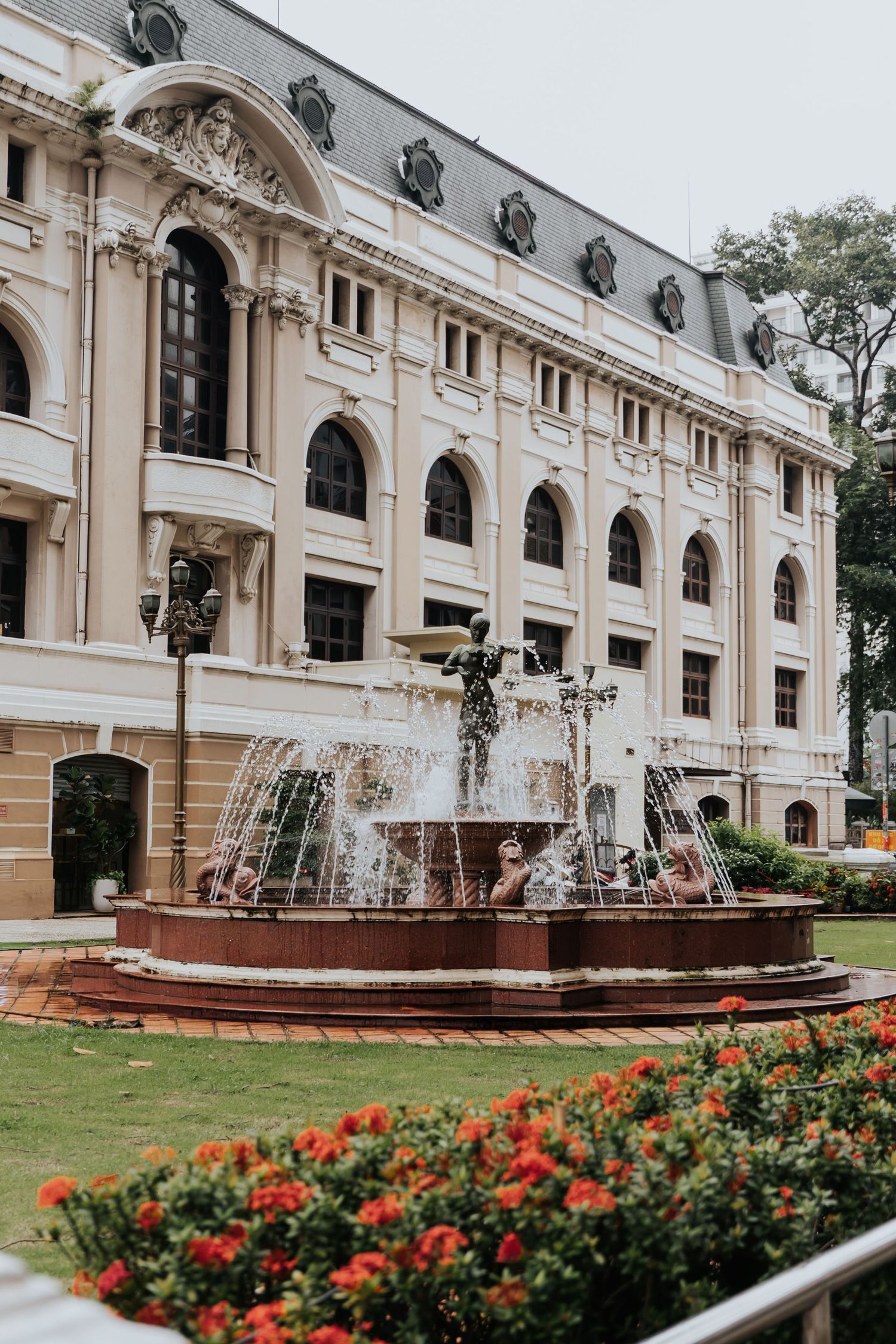 Ornate French colonial building with decorative fountain and red flowers in foreground, Ho Chi Minh City, Vietnam.
