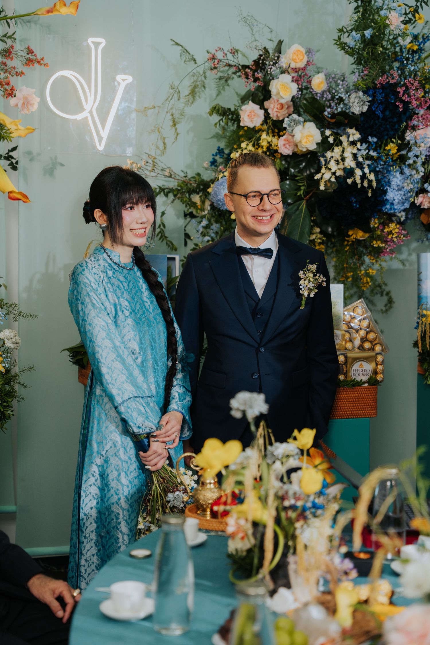 Couple in blue kebaya and navy suit pose before floral backdrop with JV monogram at elegant wedding reception.