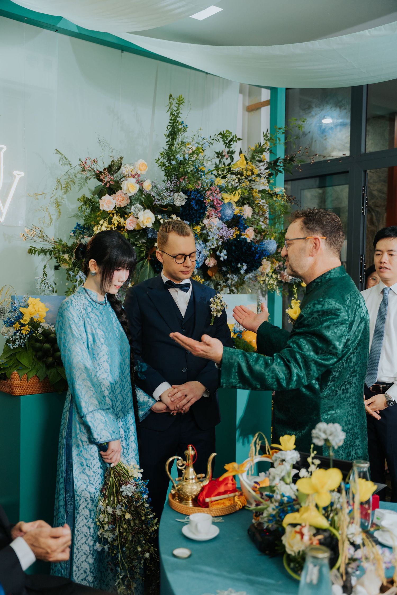 A wedding ceremony with a bride in blue ao dai, groom in suit, and elder in green traditional attire exchanging blessings.
