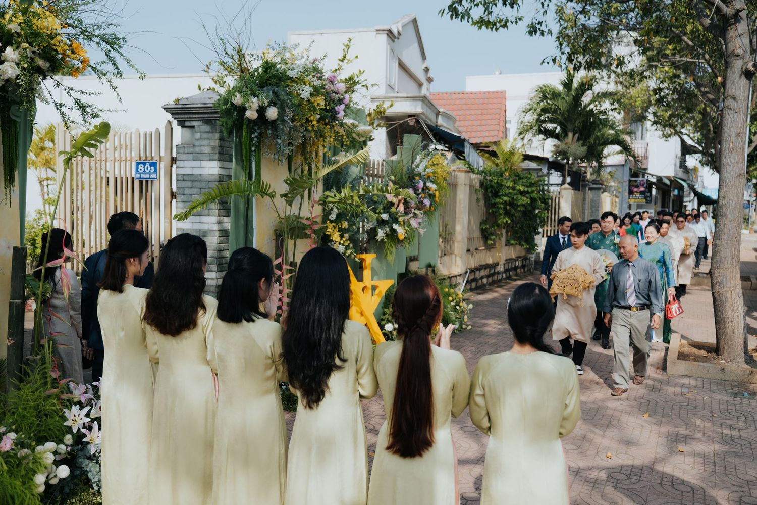 Vietnamese wedding procession with women in white ao dai traditional dresses walking toward floral arch entrance.