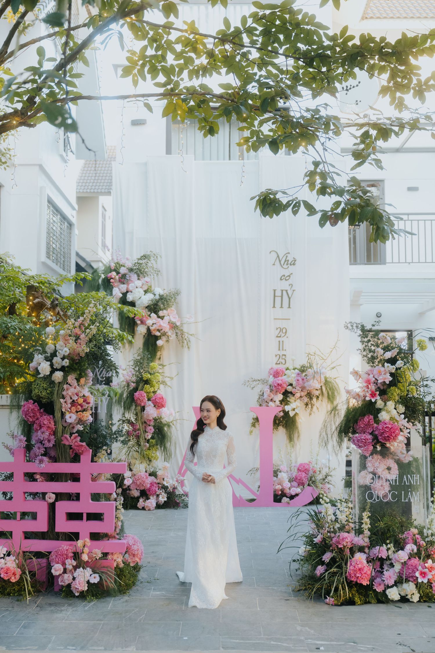 Woman in white ao dai poses at a floral Vietnamese wedding backdrop with pink flowers and decorative signage.
