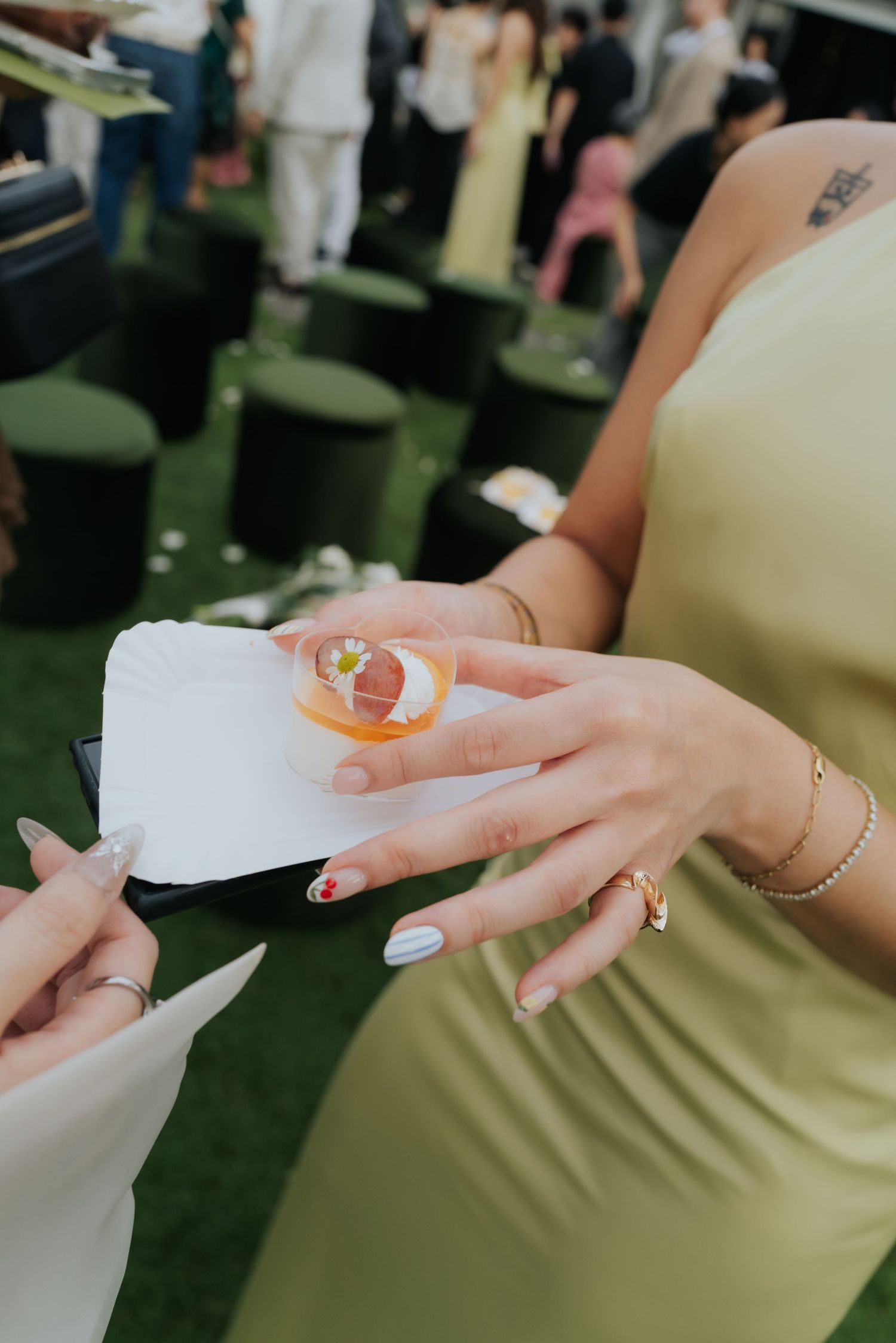 Woman in green dress holding cocktail on napkin at outdoor event, showing gold jewelry and colorful manicure.