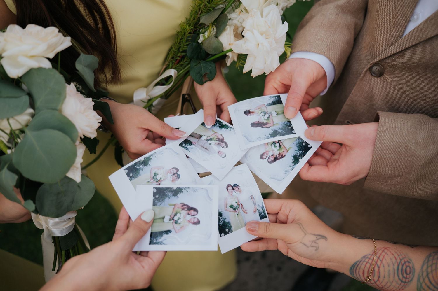 People holding Polaroid wedding photos in a circle, surrounded by white roses and eucalyptus bouquets at a wedding celebration.