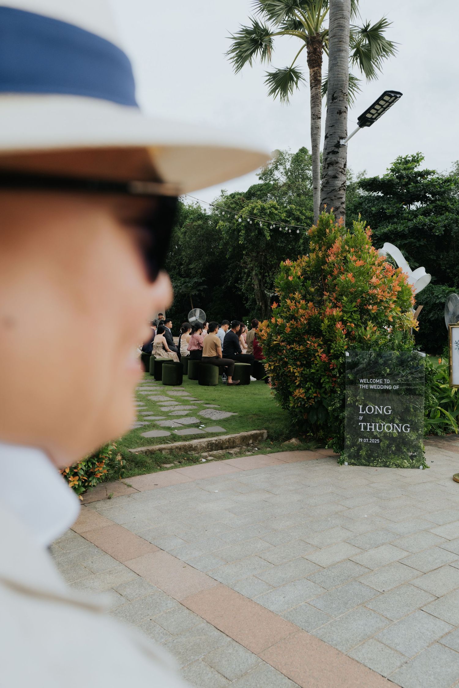 Outdoor wedding ceremony with guests seated in a garden, a welcome sign reading Long and Thuong, palm trees, and a man in a white hat.